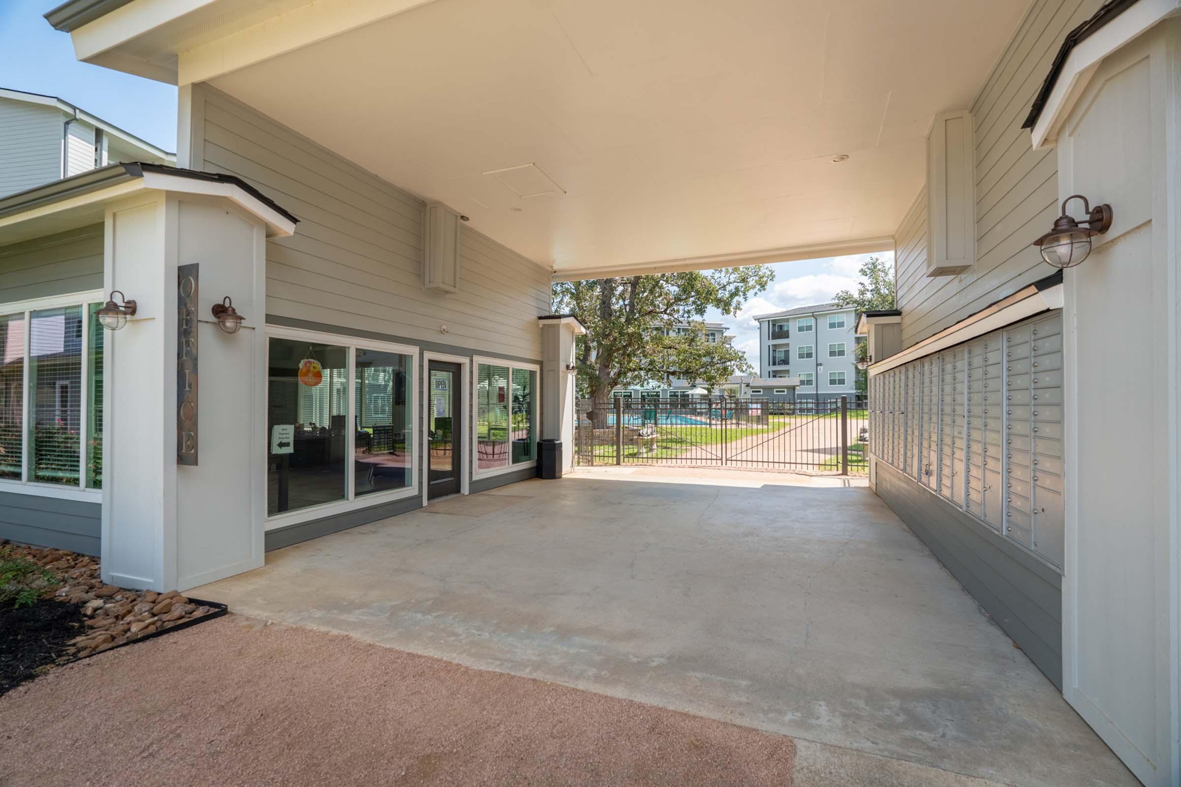 View of a covered entryway leading into a residential building. The entrance features large windows on one side, with a mailbox area and a clean, open space. In the background, there is a glimpse of a gated outdoor area and trees, suggesting a community atmosphere.