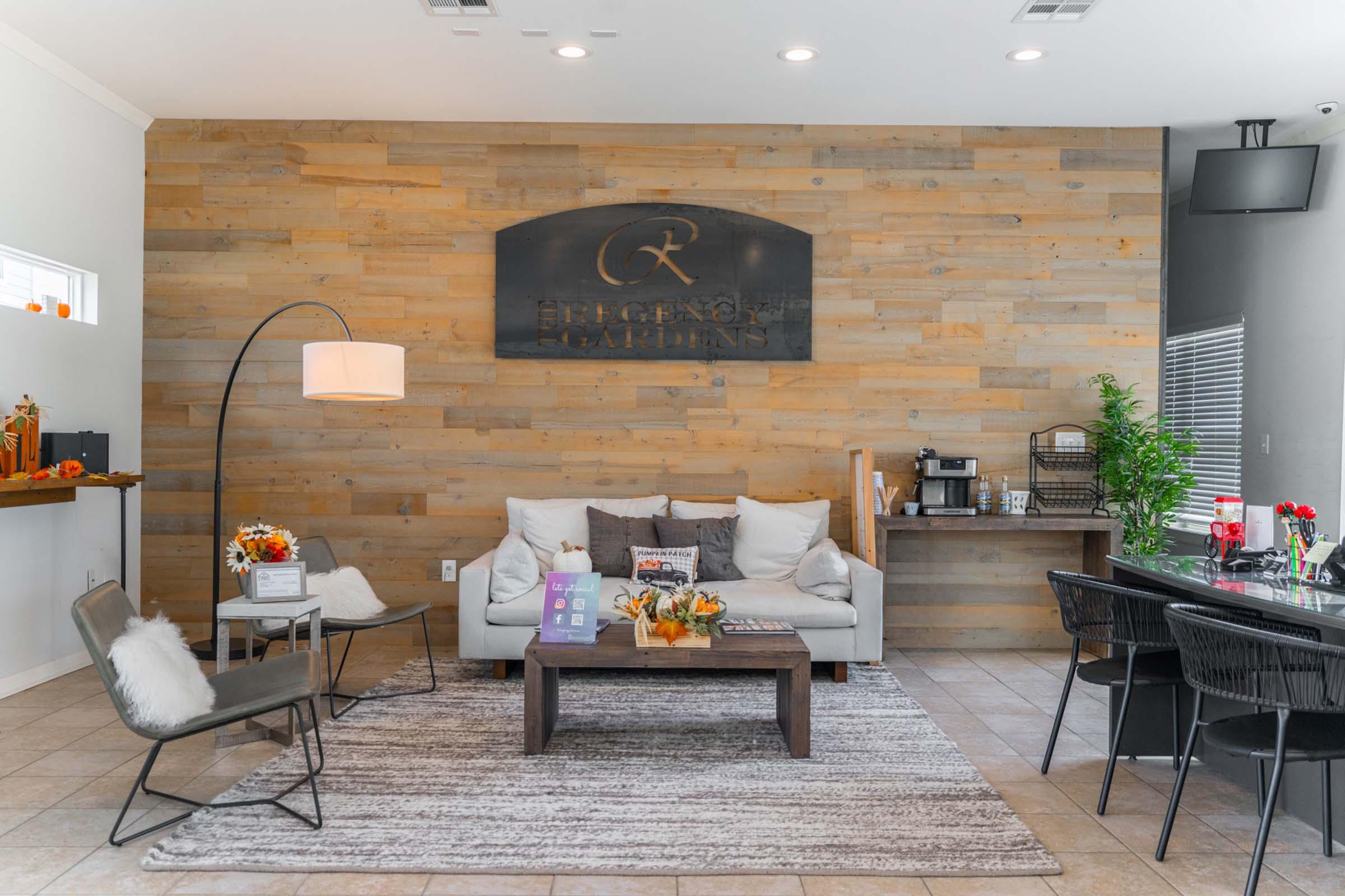 A stylish, modern reception area featuring a wooden accent wall with a sign that reads "Eugene Gardens." There is a cozy seating arrangement with a light-colored sofa, decorative pillows, and a coffee table. A floor lamp and potted plants add warmth, while a dining table with chairs is positioned alongside.