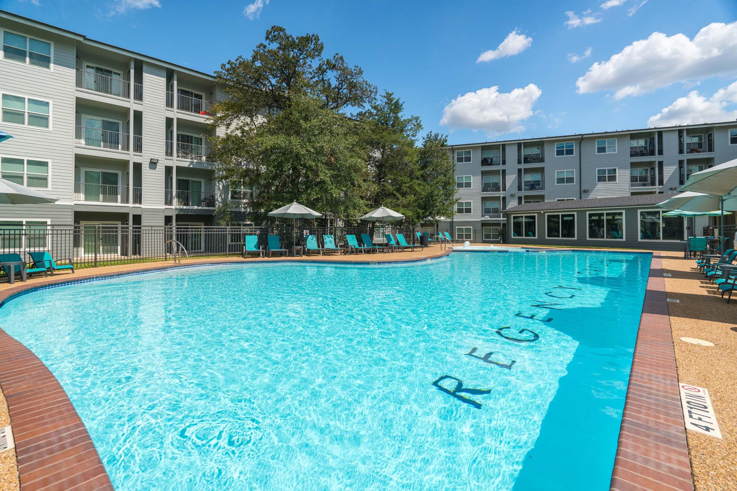 A sunny outdoor swimming pool surrounded by lounge chairs and umbrellas, with a backdrop of modern apartment buildings. The pool has the word "REGENT" clearly visible on its bottom. Trees provide some shade, and a clear blue sky completes the serene scene.