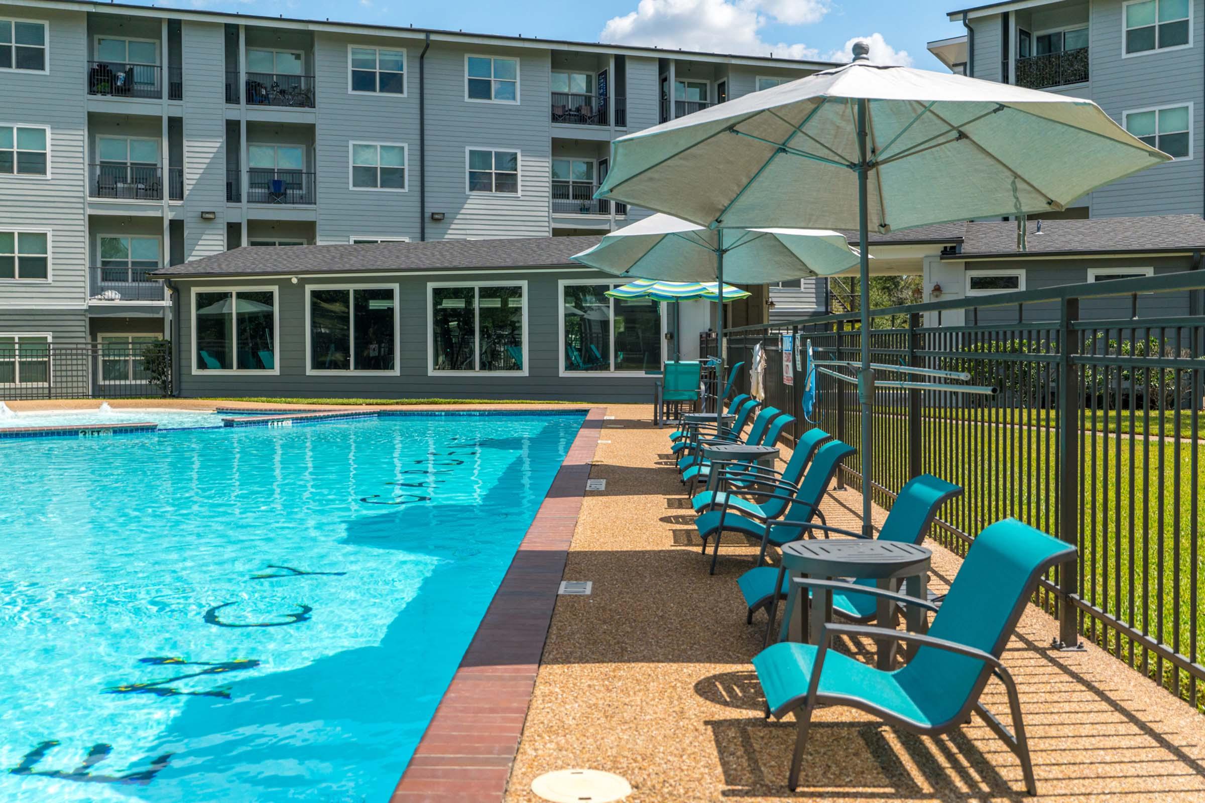 A sparkling blue swimming pool surrounded by lounge chairs and umbrellas, with a grassy area nearby. The background features a modern, multi-story building with balconies. The scene is bright and inviting, perfect for relaxation and leisure activities.