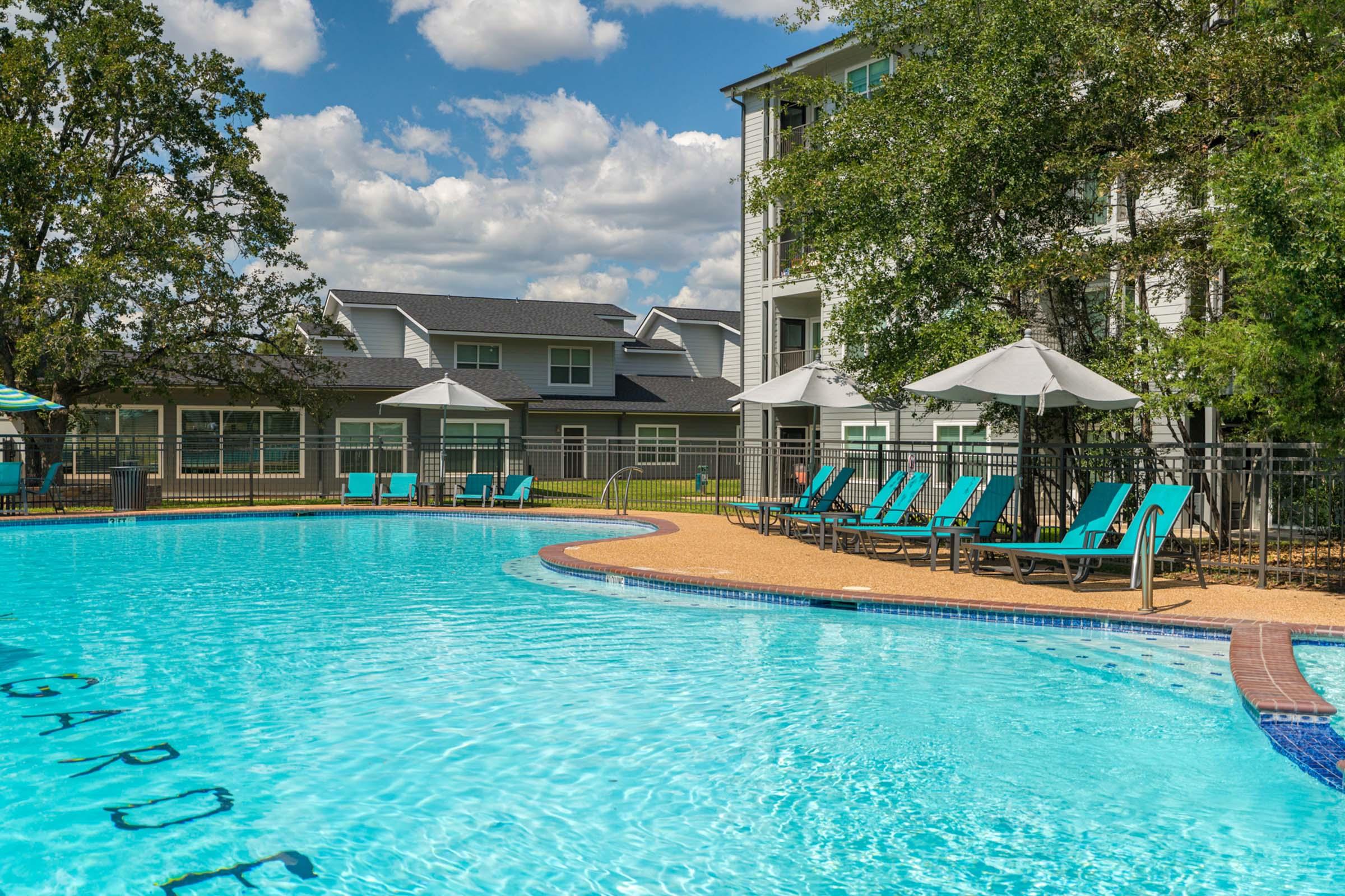 A clear blue swimming pool surrounded by lounge chairs and umbrellas, with green trees in the background. Residential buildings are visible nearby, under a bright sky with fluffy white clouds. The area looks inviting and well-maintained, perfect for relaxation.