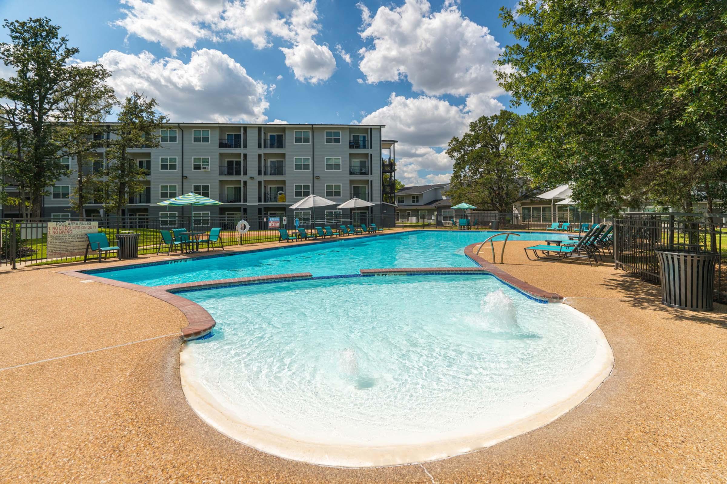A sunny pool area featuring clear blue water, lounge chairs, and umbrellas. Surrounding the pool are well-maintained lawns and trees, with a multi-story residential building in the background. The sky is filled with fluffy clouds, creating a relaxing outdoor environment.