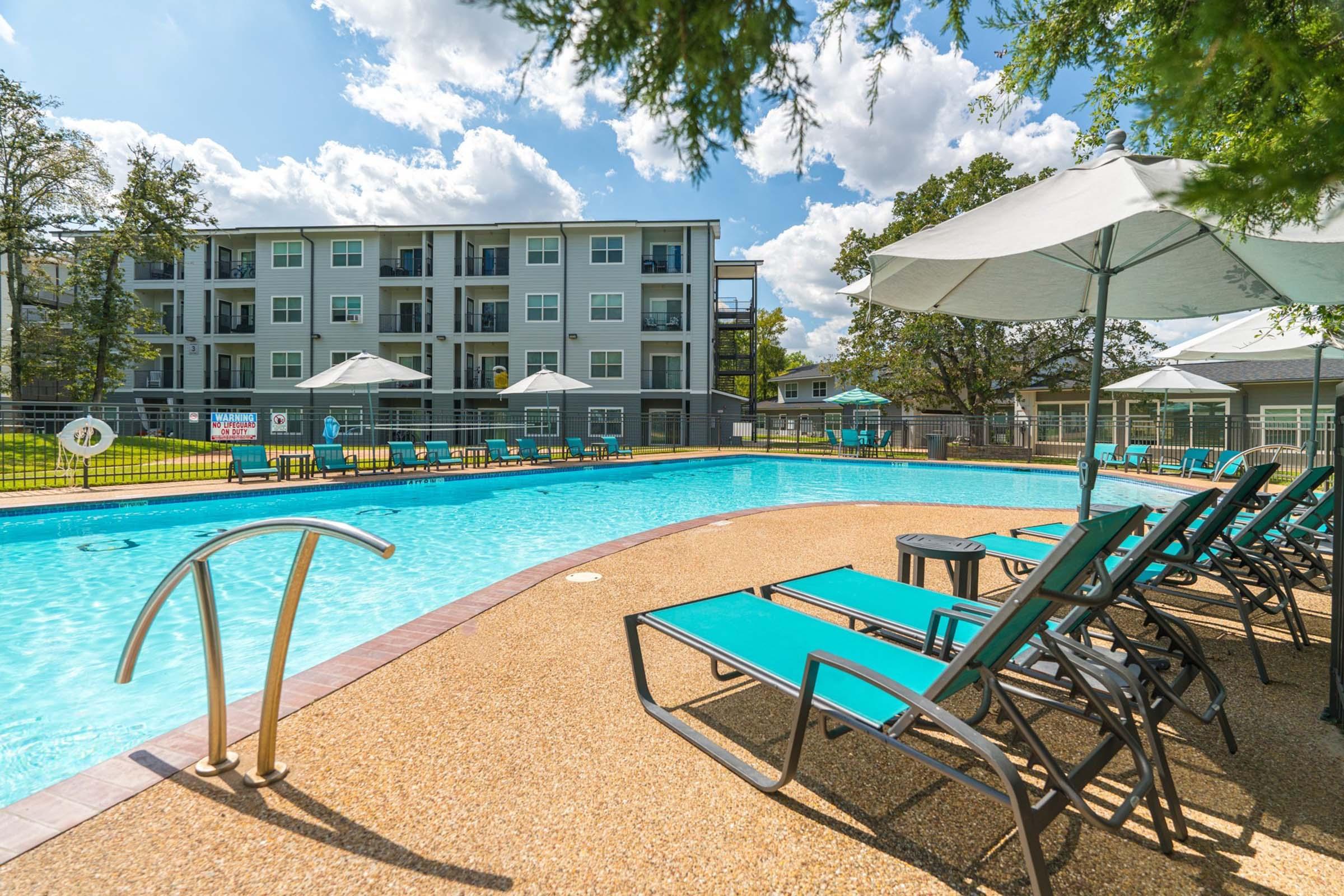 A sparkling outdoor swimming pool surrounded by lounge chairs and large umbrellas, with modern apartment buildings in the background. The scene features clear blue skies and lush greenery, creating a relaxing atmosphere for residents and guests.