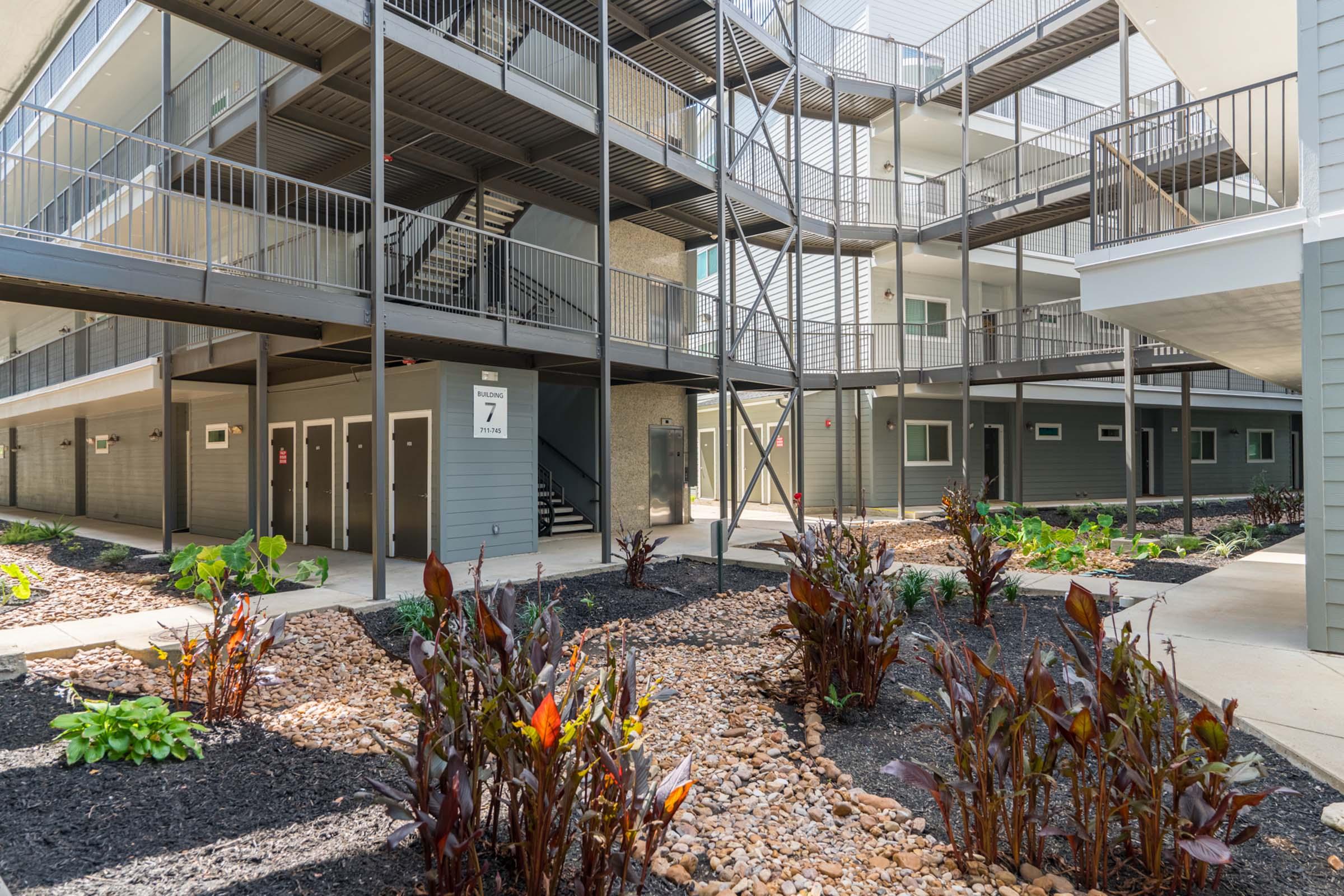 A modern apartment complex courtyard featuring multiple levels of balconies with metal railings. The area includes lush vegetation, with plants and decorative gravel paths, creating a welcoming outdoor space amidst the buildings.