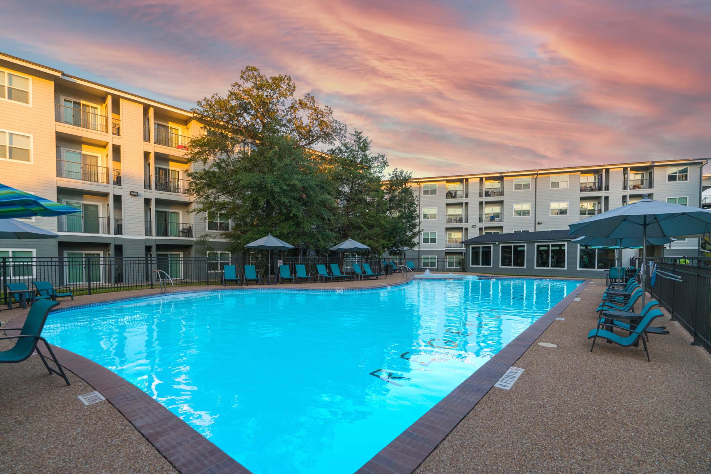 A serene swimming pool surrounded by lounge chairs and umbrellas, situated in a residential complex. The pool reflects a vibrant sunset sky, with multi-story buildings visible in the background. The scene conveys a relaxing outdoor atmosphere.