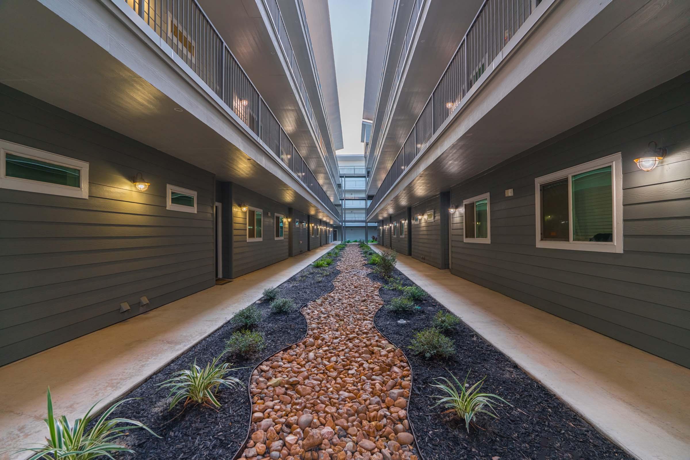 A view looking down a modern hotel corridor with two levels of rooms on either side. The pathway features smooth pebbles and small plants, creating a serene atmosphere with soft lighting illuminating the area. The building has a contemporary design with gray siding and glass panels.