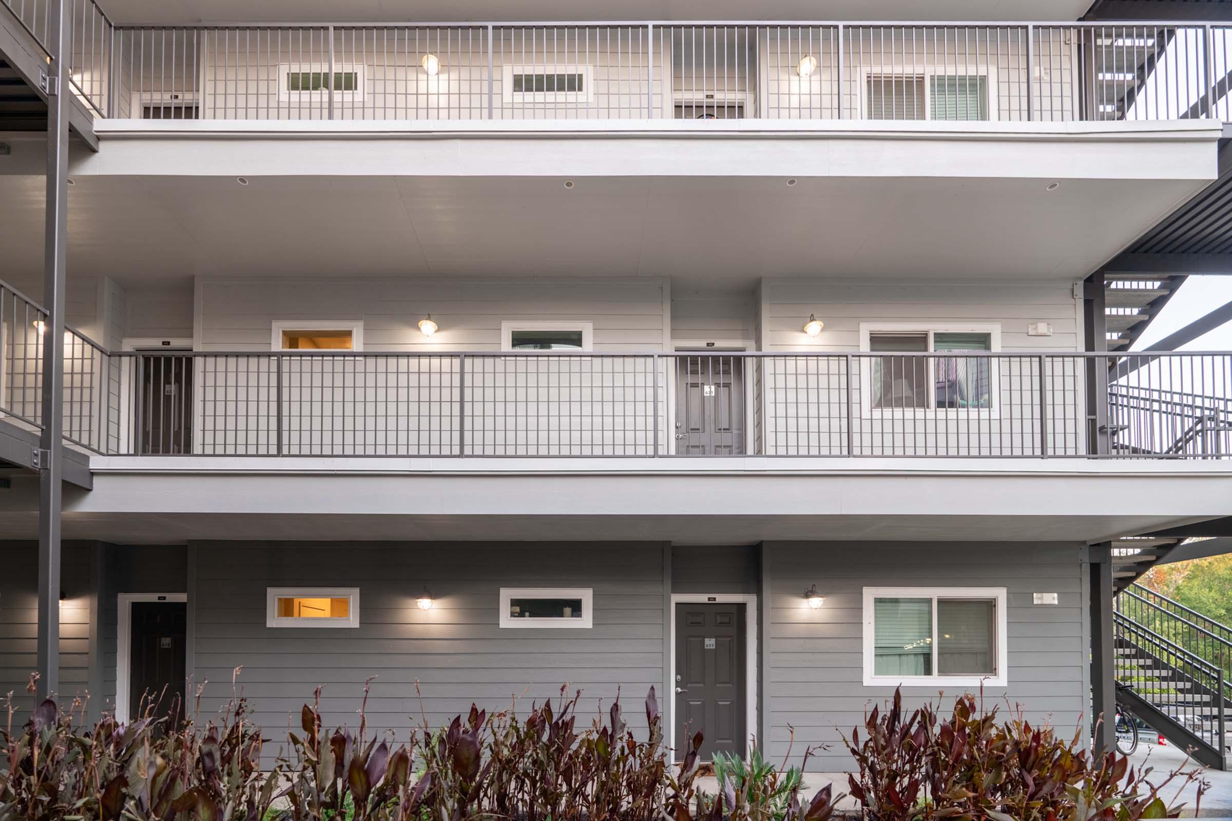 Exterior view of a two-story apartment building with gray siding. Each floor features balconies with railings and doors. The lower level has two units visible, and the area is surrounded by decorative plants. Soft lighting illuminates the entrances, creating a welcoming atmosphere.