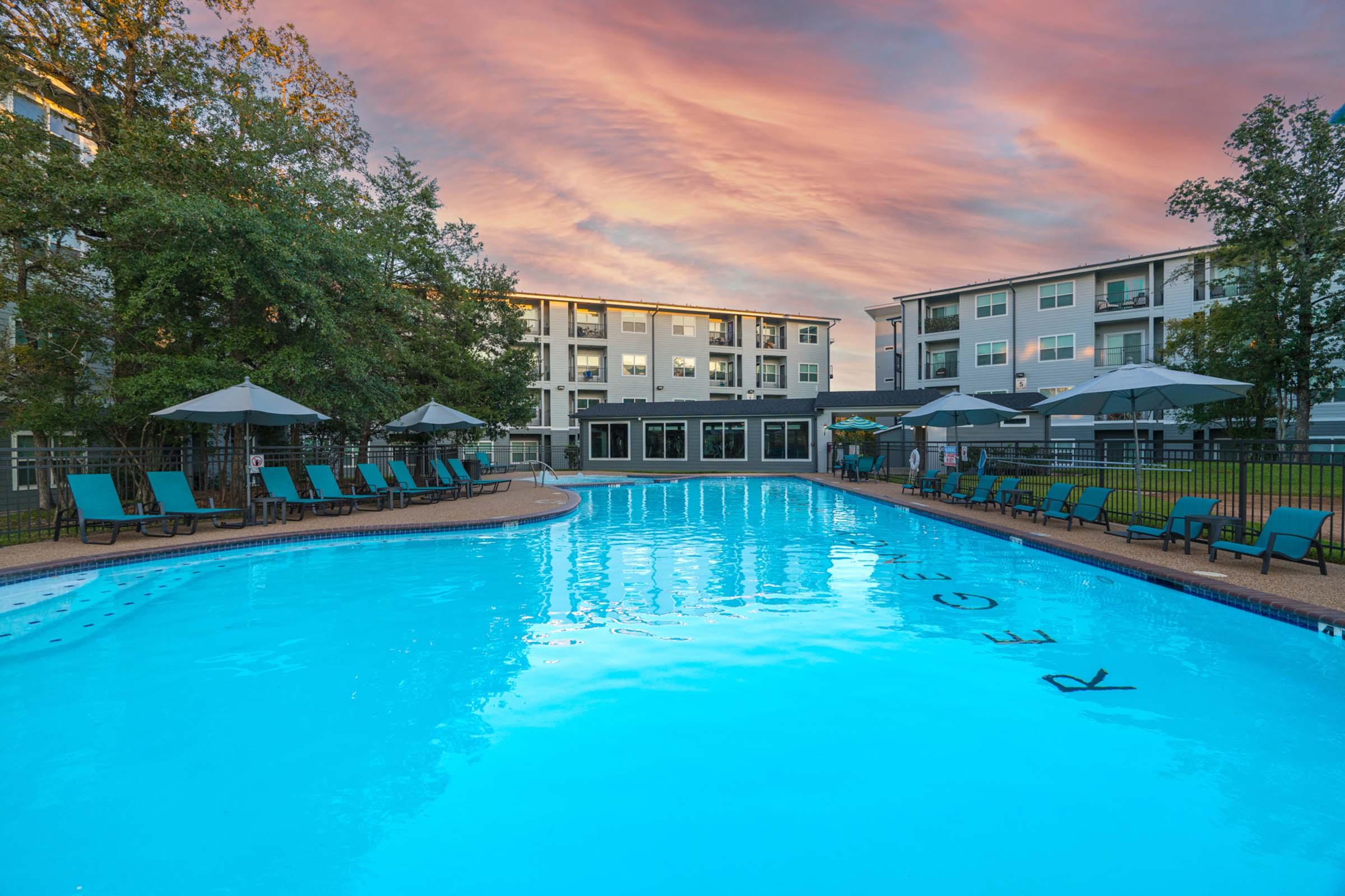 A tranquil swimming pool surrounded by lounge chairs and umbrellas, with a modern apartment building in the background. The sky is adorned with vivid pink and orange hues, creating a serene atmosphere.