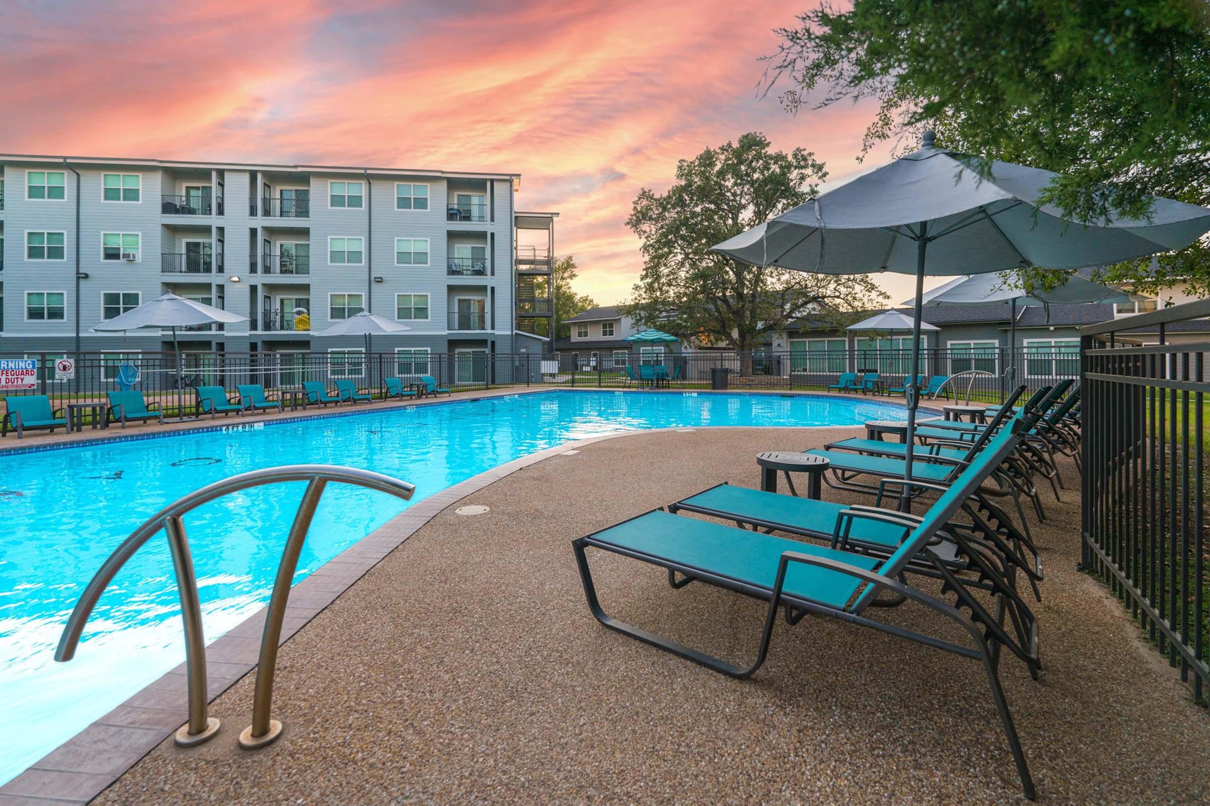 A serene swimming pool surrounded by lounge chairs and umbrellas, with a multi-story building in the background. The sky is painted with vibrant sunset colors, adding a warm ambiance to the scene. The pool area is well-maintained, inviting for relaxation and leisure activities.