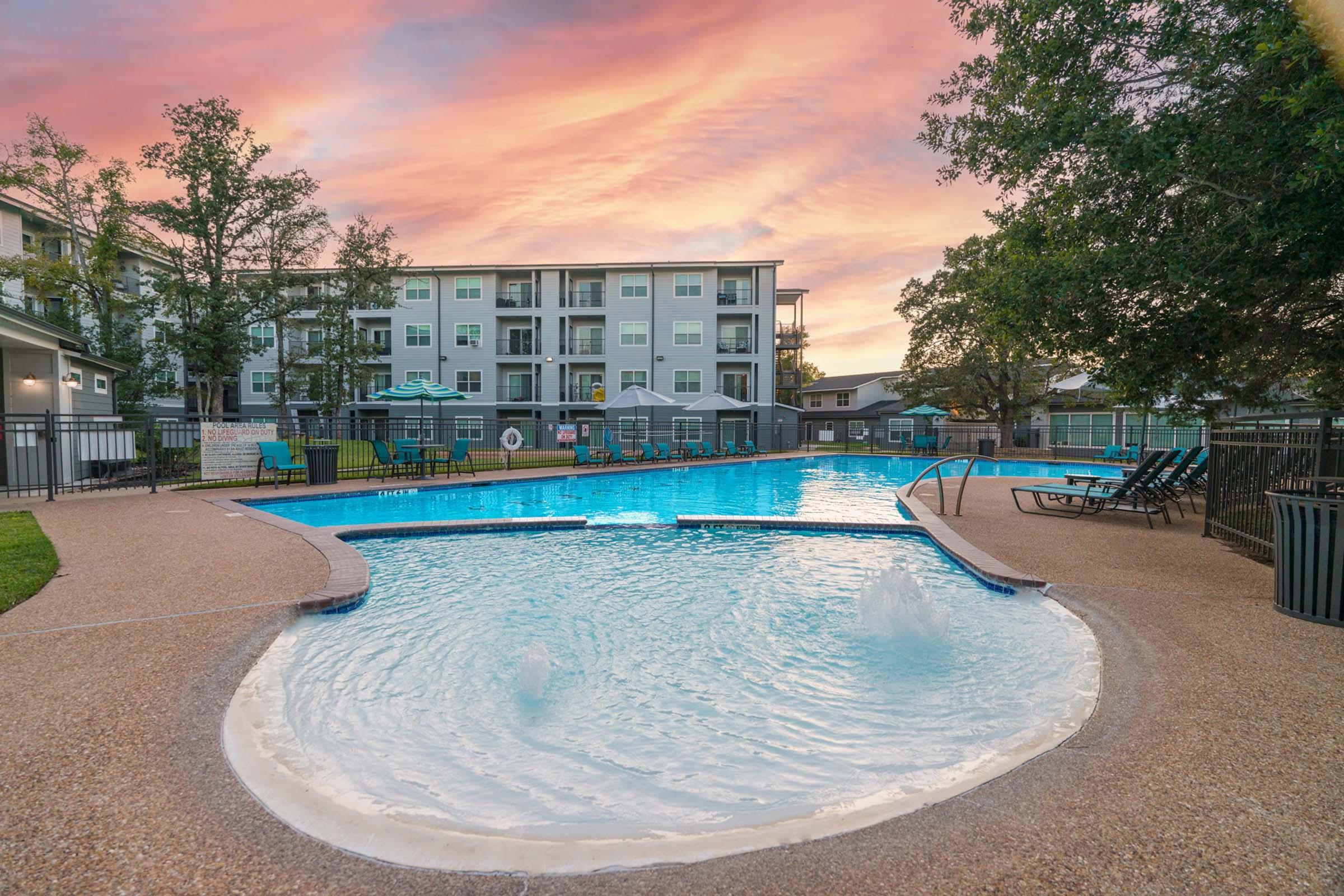 A serene swimming pool surrounded by lounge chairs and vibrant landscaping, with a backdrop of an apartment complex under a colorful sunset sky. The pool features gentle water fountains, creating a relaxing atmosphere for residents and guests.