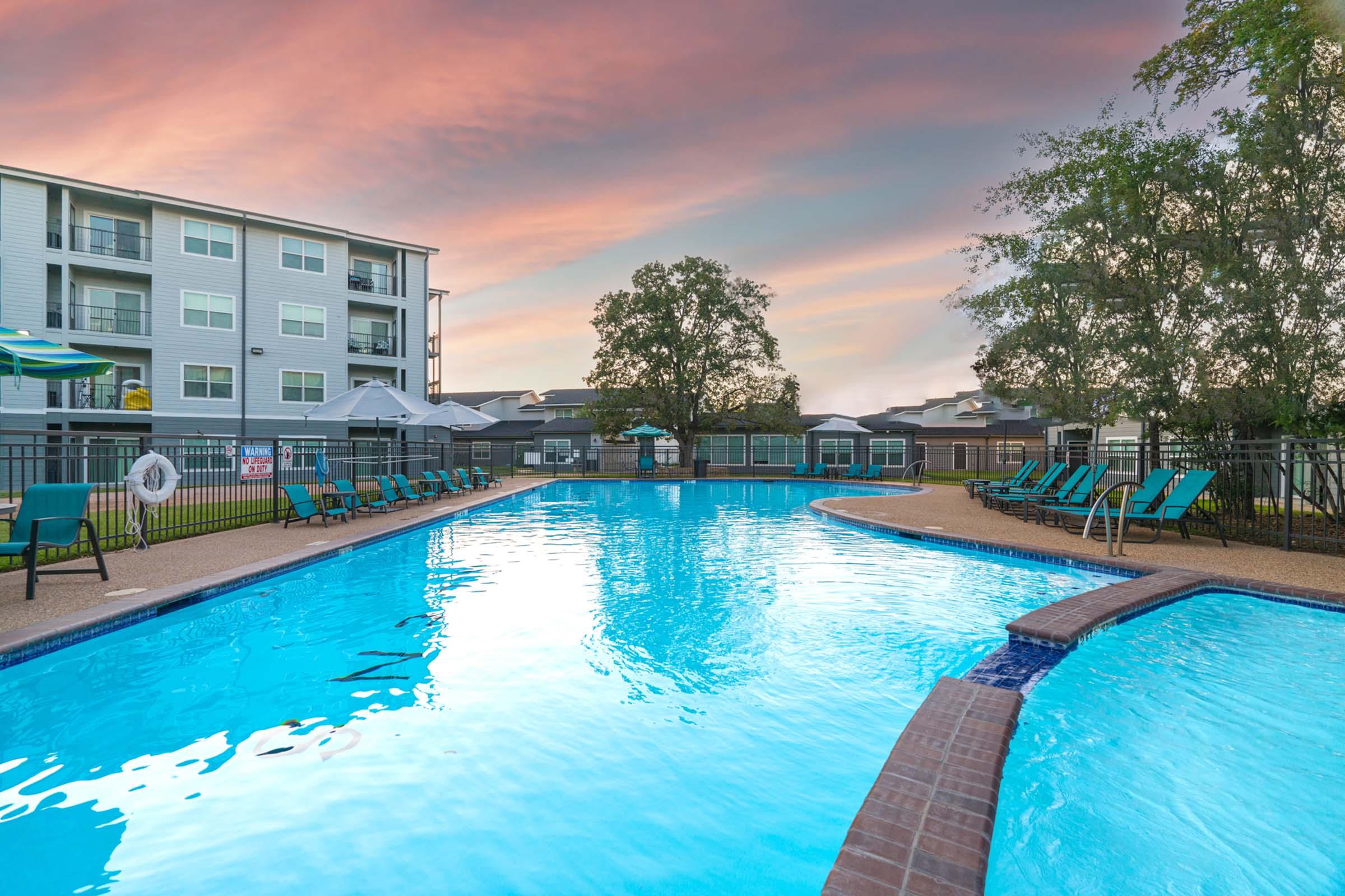A swimming pool surrounded by lounge chairs under a colorful sunset sky. Several umbrellas and trees are visible in the background, alongside modern apartment buildings. The scene conveys a relaxing atmosphere, ideal for leisure and recreation.
