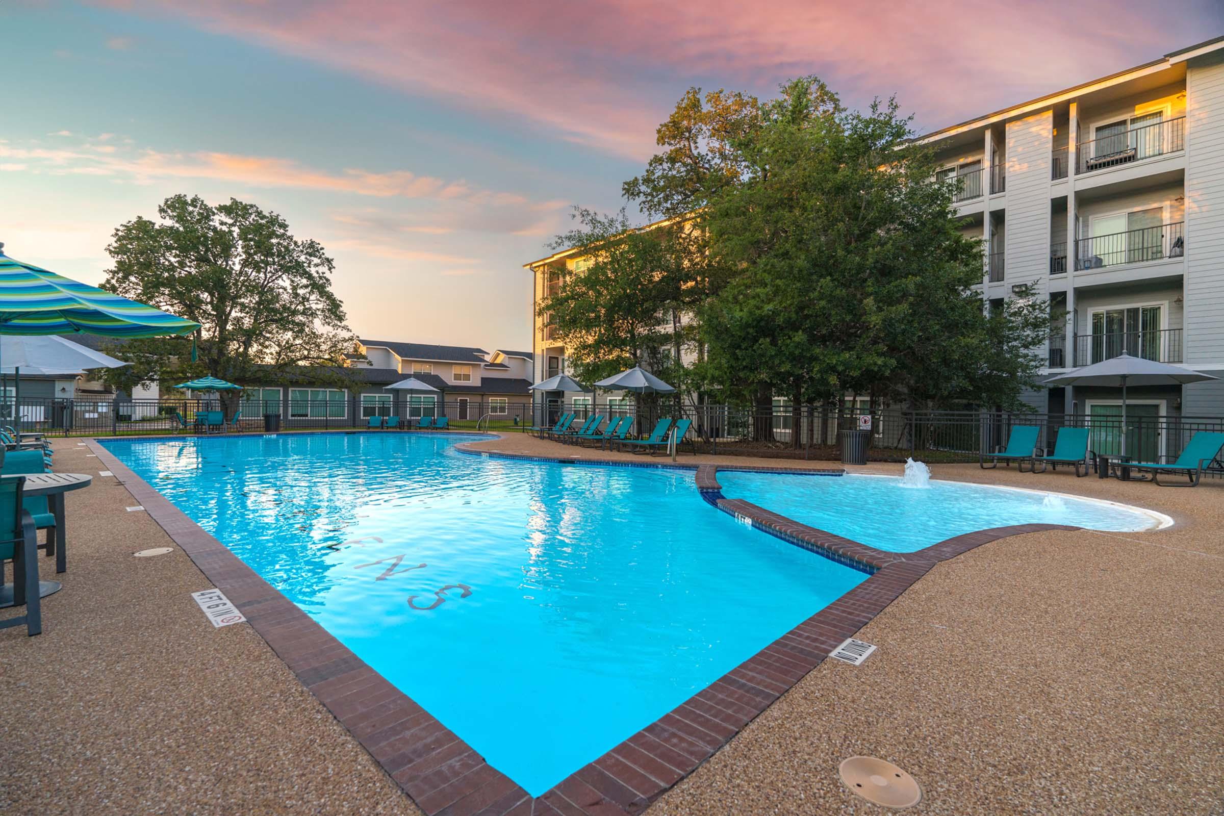 A scenic swimming pool surrounded by lounge chairs and umbrellas, set against a backdrop of a modern building. The water is a bright blue, reflecting the colorful sky during sunset. Green trees add a touch of nature to the serene environment, creating a relaxing atmosphere for guests.