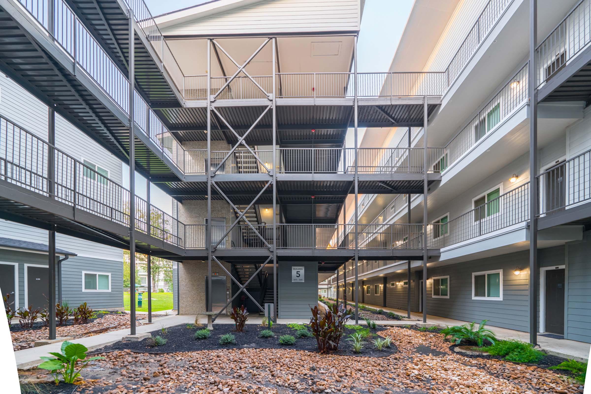 A view of an apartment complex with multiple floors and metal staircases. The courtyard features landscaped areas with plants and gravel pathways. Natural light highlights the modern building design, showcasing a combination of grey and white exterior finishes.