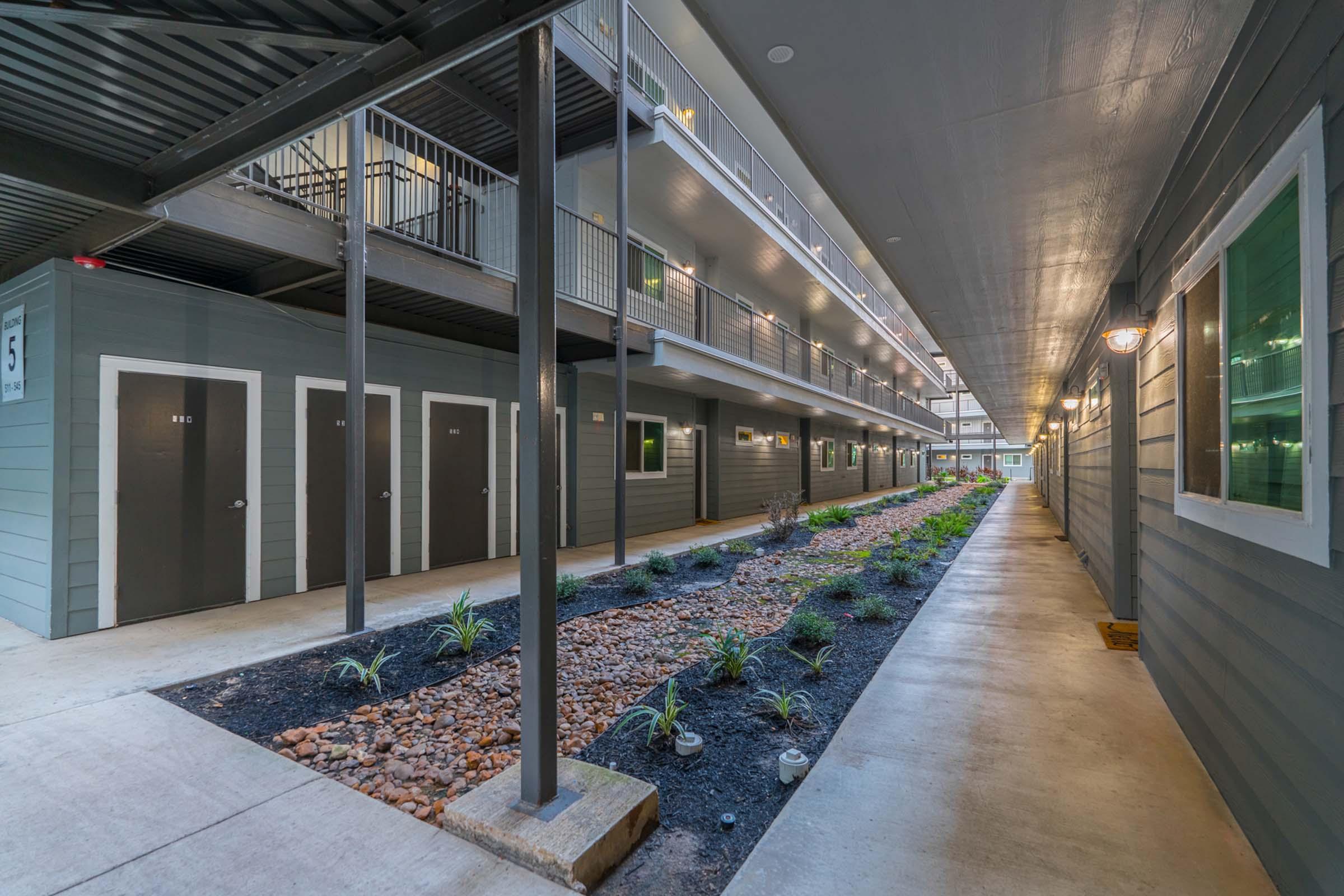 A corridor in an apartment building featuring multiple doors on either side, with a landscaped area in the center. The pathway is lined with small plants and decorative rocks, and the building has a modern design with gray panels and white trim. Soft lighting illuminates the space.
