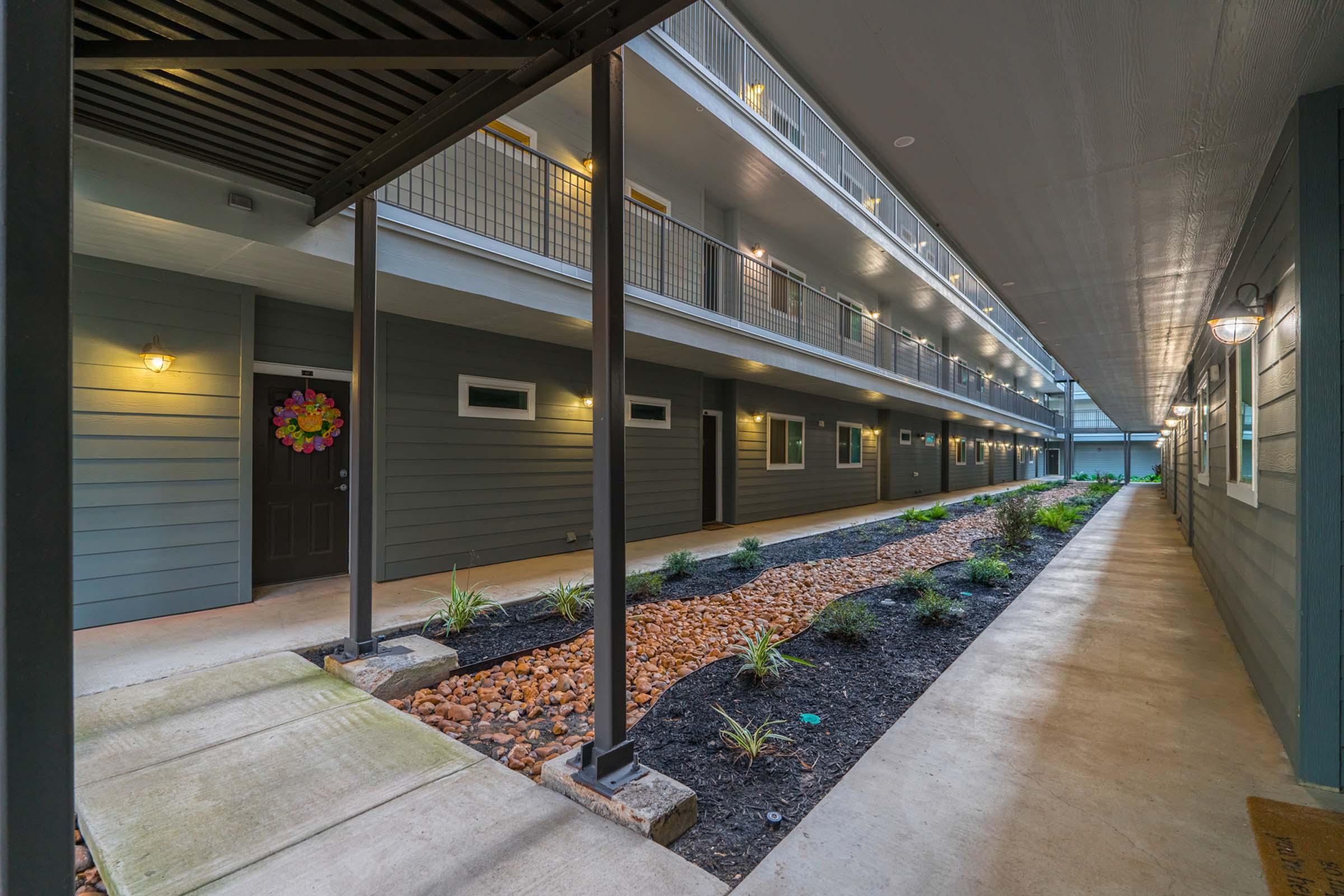 View of an interior hallway in an apartment building, featuring gray exterior walls, lit doorways of individual units, and a landscaped area with small plants and decorative stones along the ground. The corridor is well-lit, showcasing a modern design with clean lines and a welcoming atmosphere.