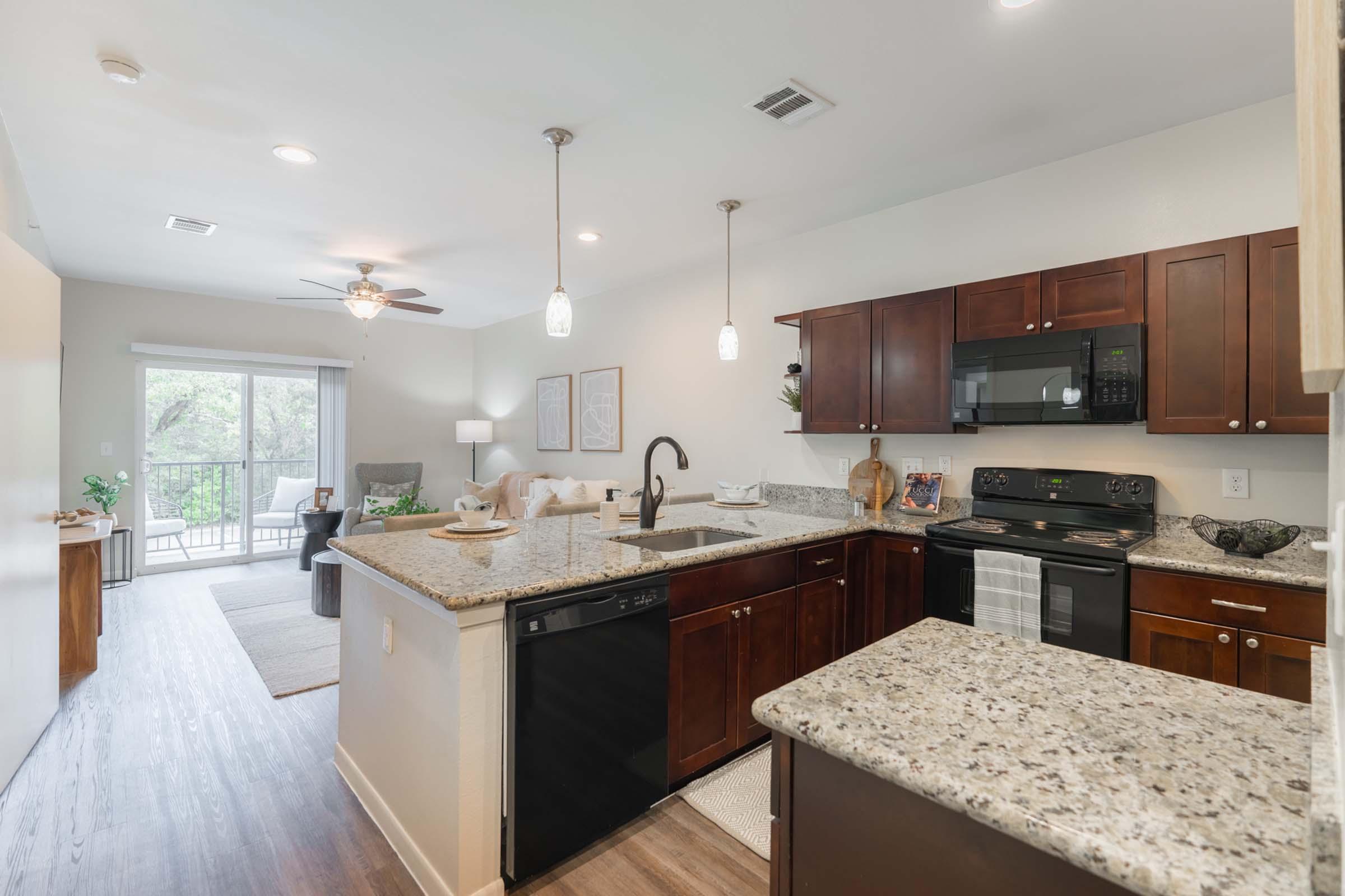 A modern kitchen with dark wood cabinetry, granite countertops, and stainless steel appliances, including a microwave and dishwasher. The room features pendant lighting and overlooks a spacious living area with a couch and a sliding door leading to a balcony. Natural light brightens the space.