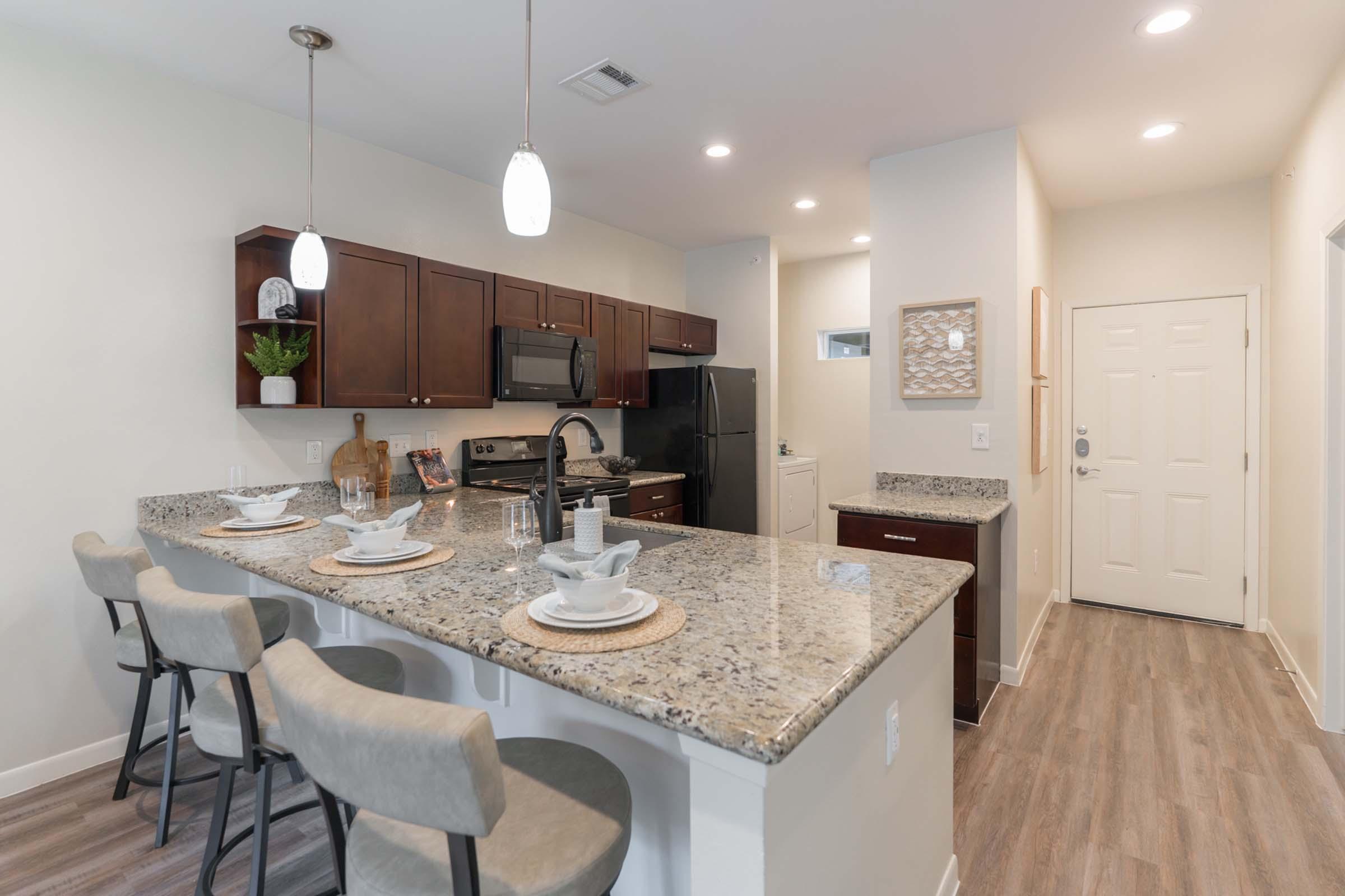 A modern kitchen featuring dark cabinetry, stainless steel appliances, and a granite countertop island with four bar stools. One side of the kitchen has an open layout leading to a door, while the other includes a small sink area. Soft lighting and neutral colors create a warm and inviting atmosphere.