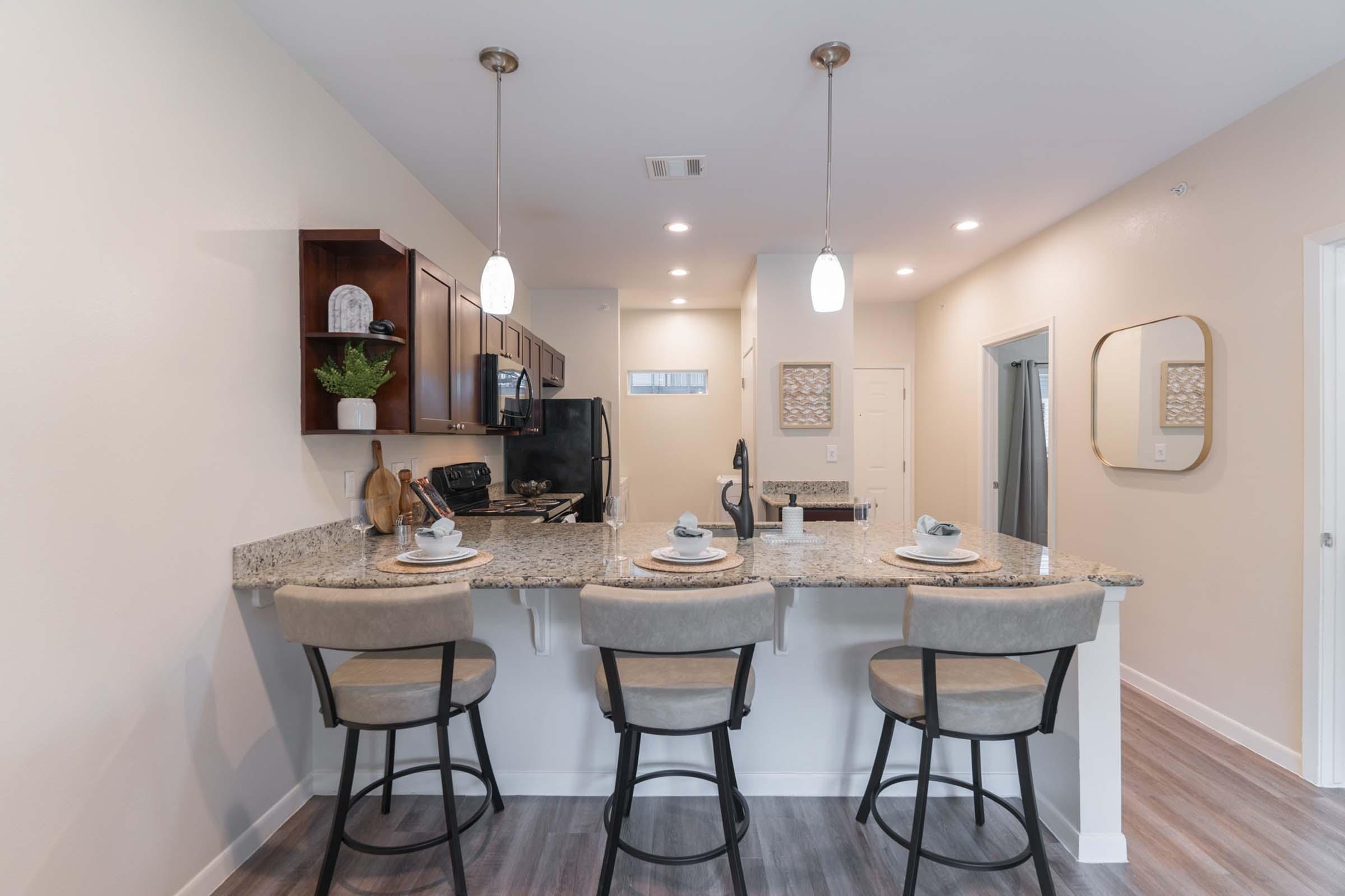 A modern kitchen featuring a granite countertop with three bar stools, pendant lighting, and a black refrigerator. The kitchen is equipped with wooden cabinets and looks well-organized and inviting. A small dining setup is visible, adding a cozy touch to the space.