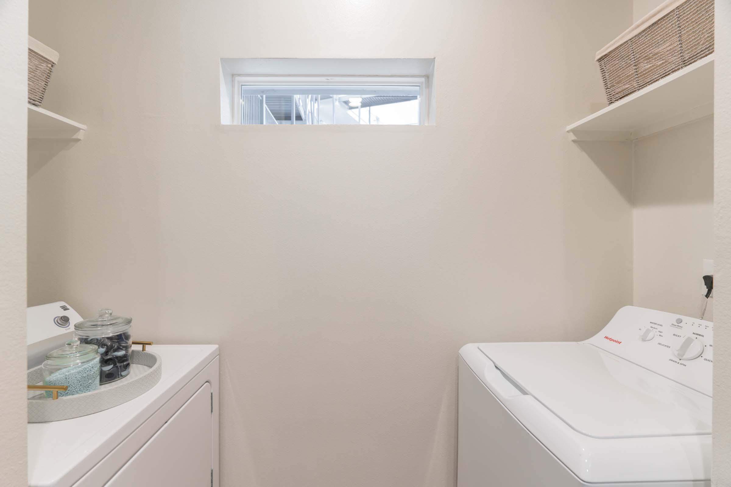 A clean laundry room featuring a white washer and dryer, with a small window for natural light. On the left, there’s a round basket containing laundry supplies. The room's walls are painted beige, and there are additional storage baskets on the upper shelves.