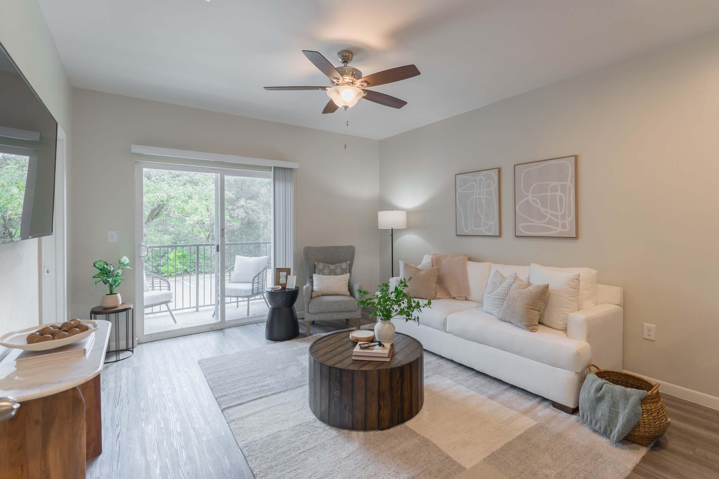 A cozy living room featuring a white sofa with decorative pillows, a wooden coffee table, and a comfortable gray armchair. There’s a ceiling fan, a lamp, and wall art on the neutral-toned walls. A balcony with outdoor seating is visible through sliding glass doors, with greenery outside providing a peaceful backdrop.