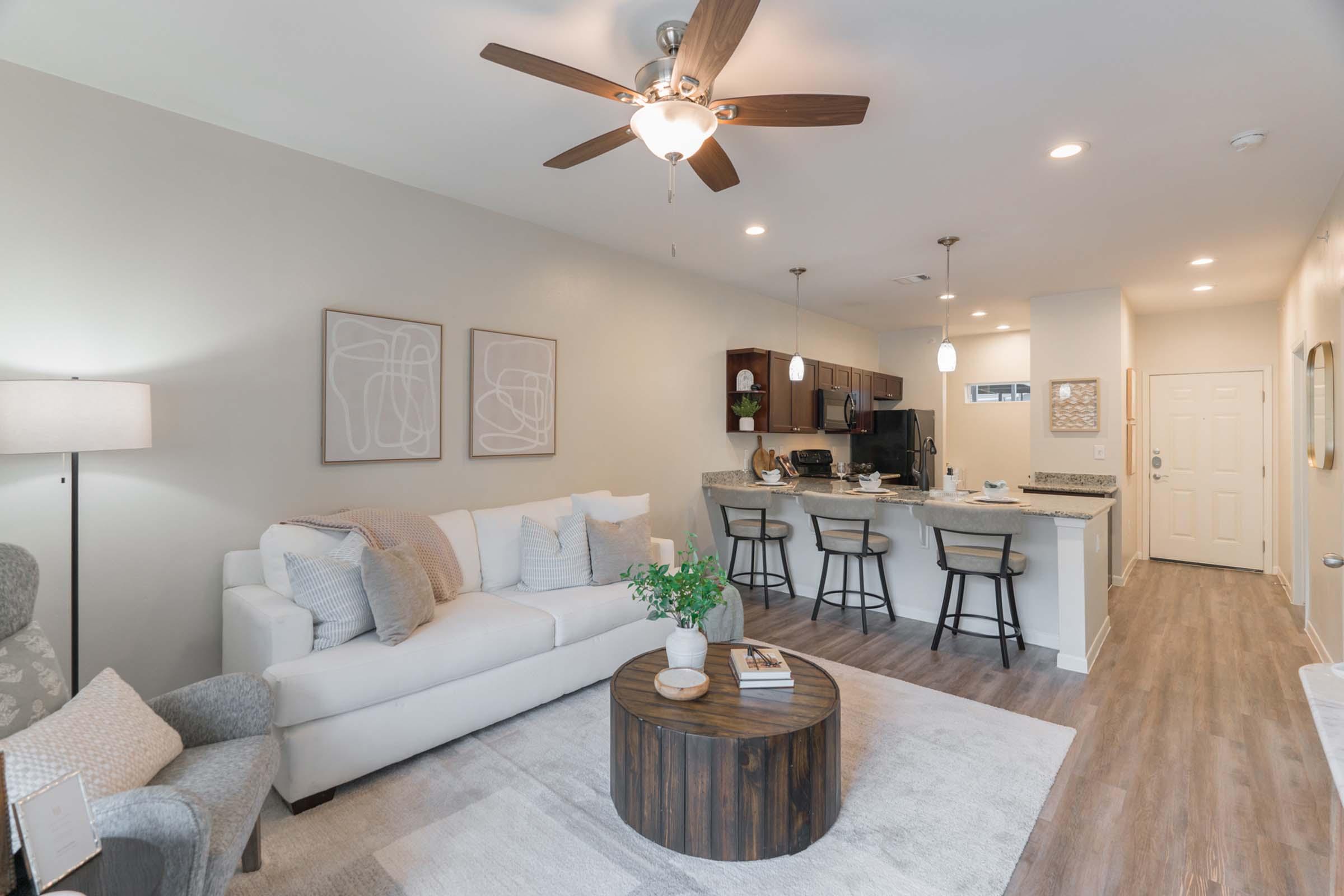 A modern, open-concept living area featuring a light gray couch with decorative pillows, a round wooden coffee table, and a stylish floor lamp. The kitchen in the background has dark cabinetry and bar stools. There are decorative wall art pieces and a light-colored rug underfoot, creating a cozy atmosphere.