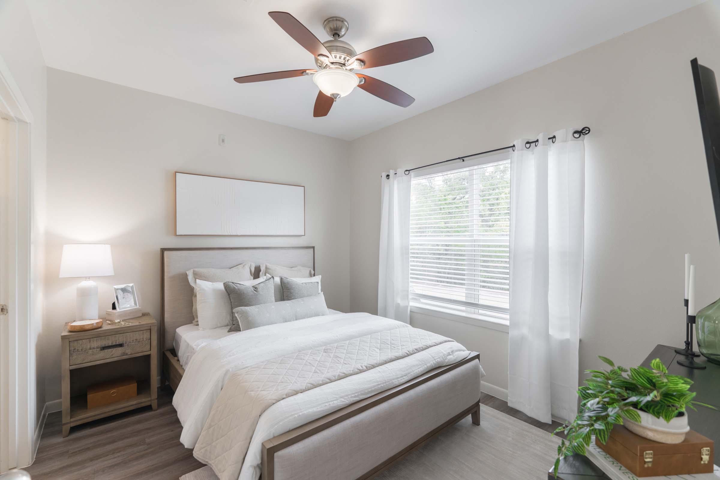 A bright and airy bedroom featuring a queen-sized bed with light bedding, a modern ceiling fan, and white curtains. A bedside table with a lamp and a small plant adds warmth. A window lets in natural light, and the walls are painted in a soft neutral color, contributing to a cozy atmosphere.
