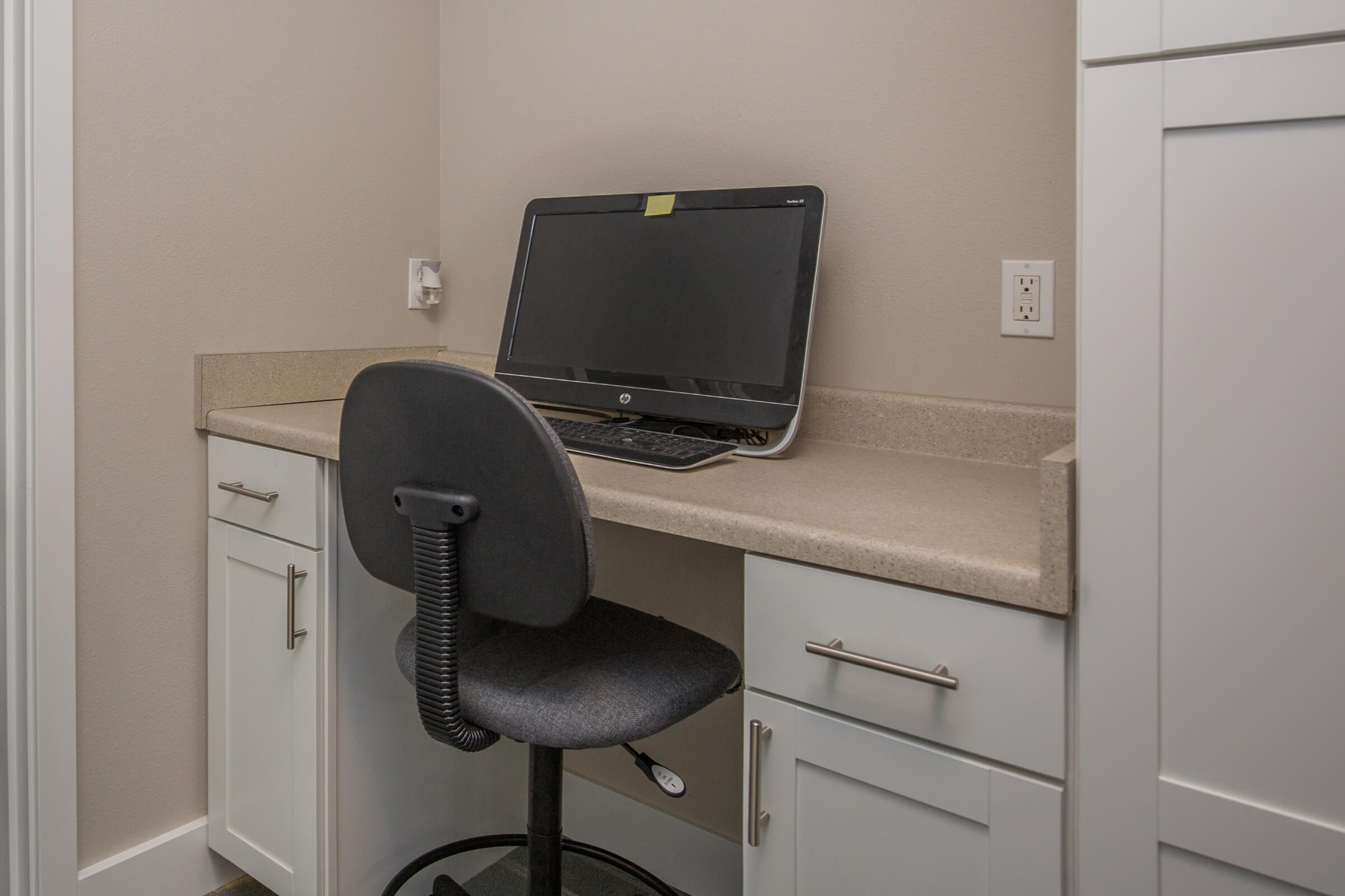 A small workspace featuring a desk with a beige countertop, a black computer monitor, and a black office chair. The desk is set against a light gray wall, with white cabinetry above and beside it. The space is organized and minimalistic, ideal for a home office or study area.
