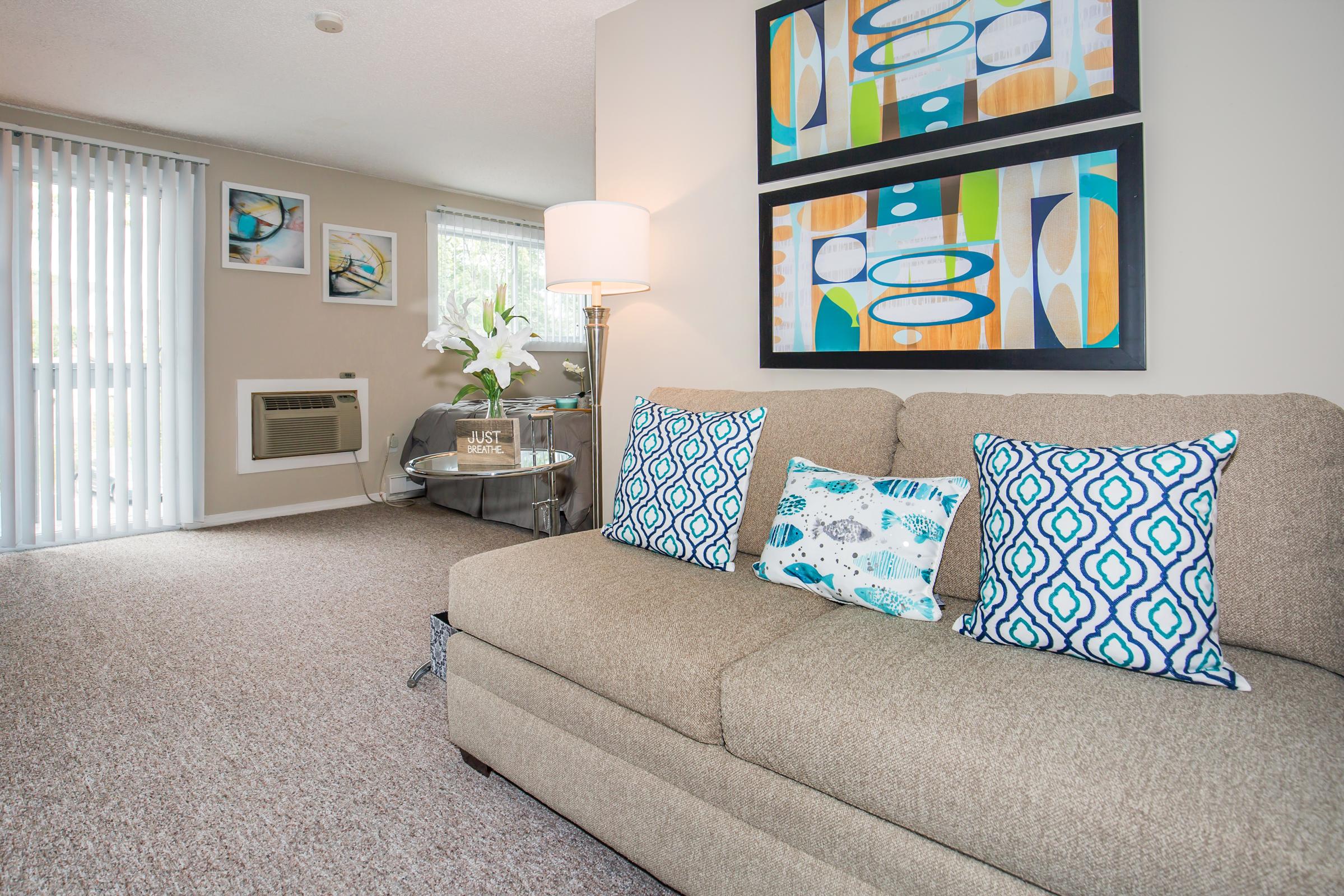 A cozy living room featuring a brown sofa with decorative pillows, two colorful framed artworks on the wall, and a lamp beside the sofa. A small table sits next to the sofa, while large windows with vertical blinds allow natural light to enter. There's an air conditioning unit visible on the wall.