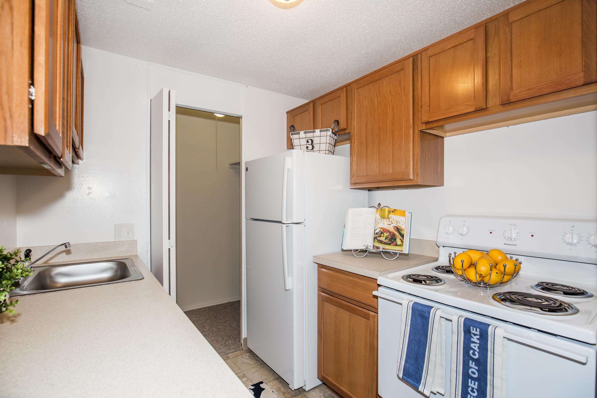 A compact kitchen featuring wooden cabinets, a white refrigerator, and an oven with a stovetop. A counter with a sink runs along one side, and a bowl of oranges sits on the counter near an open cookbook. A closet door is visible in the background, adding to the functional layout of the space.