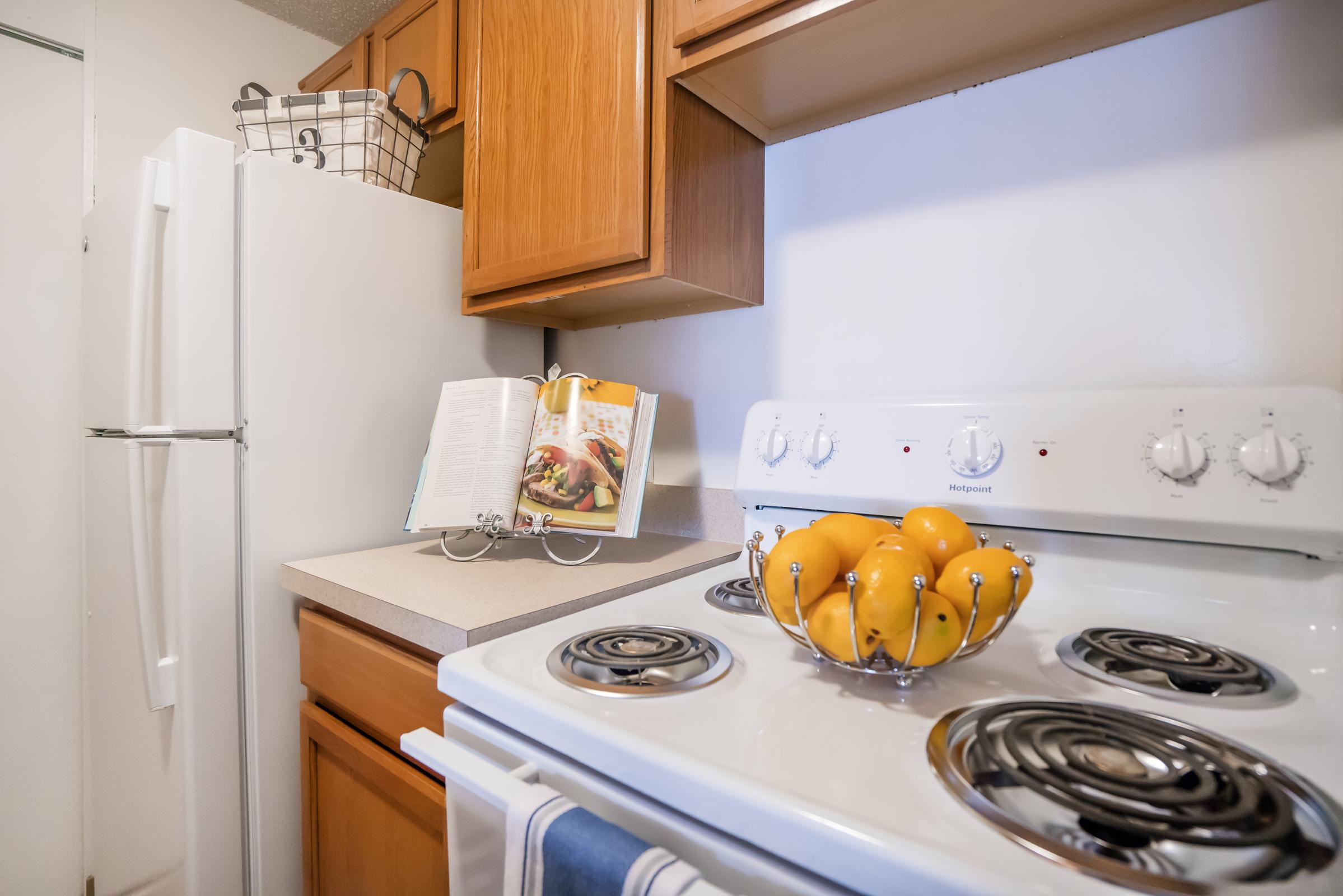 A kitchen scene featuring an open cookbook on a stand next to a white stove with knobs. A bowl of oranges is placed on the counter, and a white refrigerator is visible in the background. Brown wooden cabinets are above the counter, creating a warm and inviting cooking space.