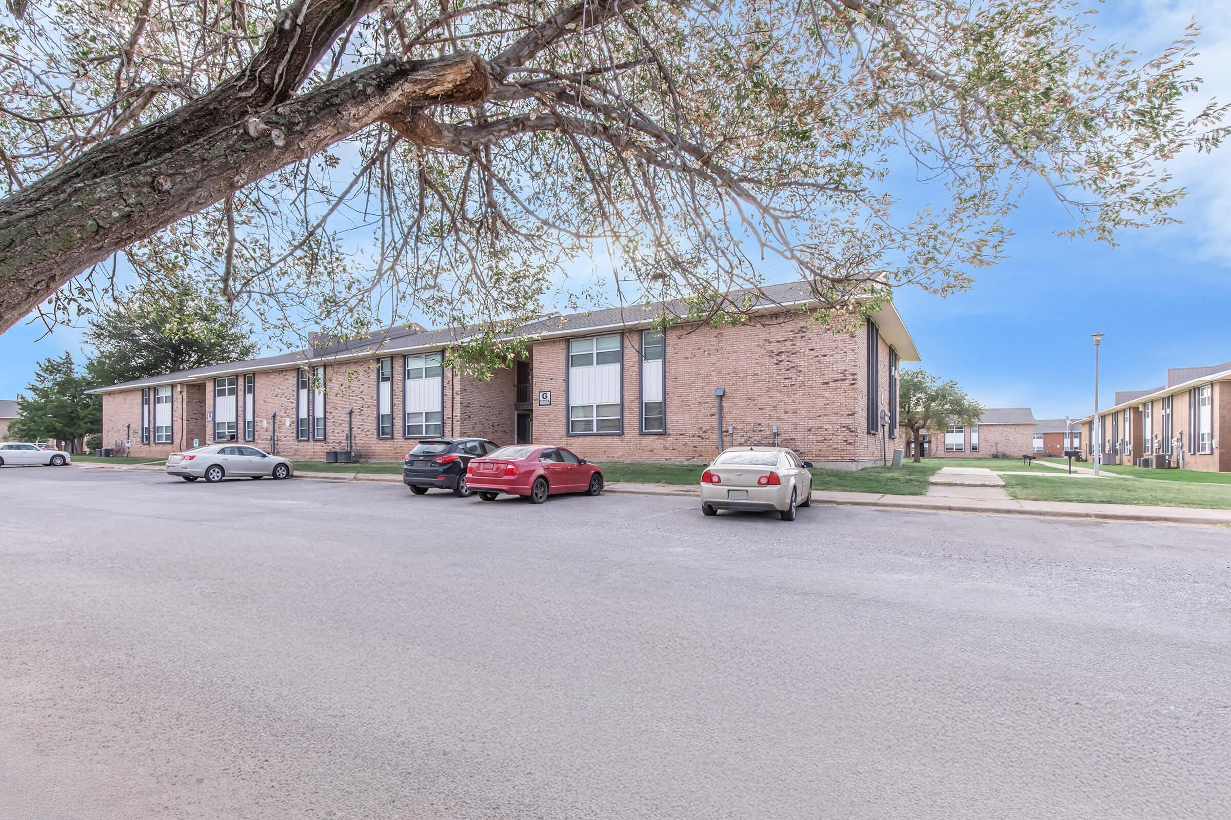 A view of an apartment complex with several cars parked in front. The two-story brick building features multiple windows and greenery around it. A large tree with sparse leaves is in the foreground, and the sky is clear. The setting appears residential and well-maintained.