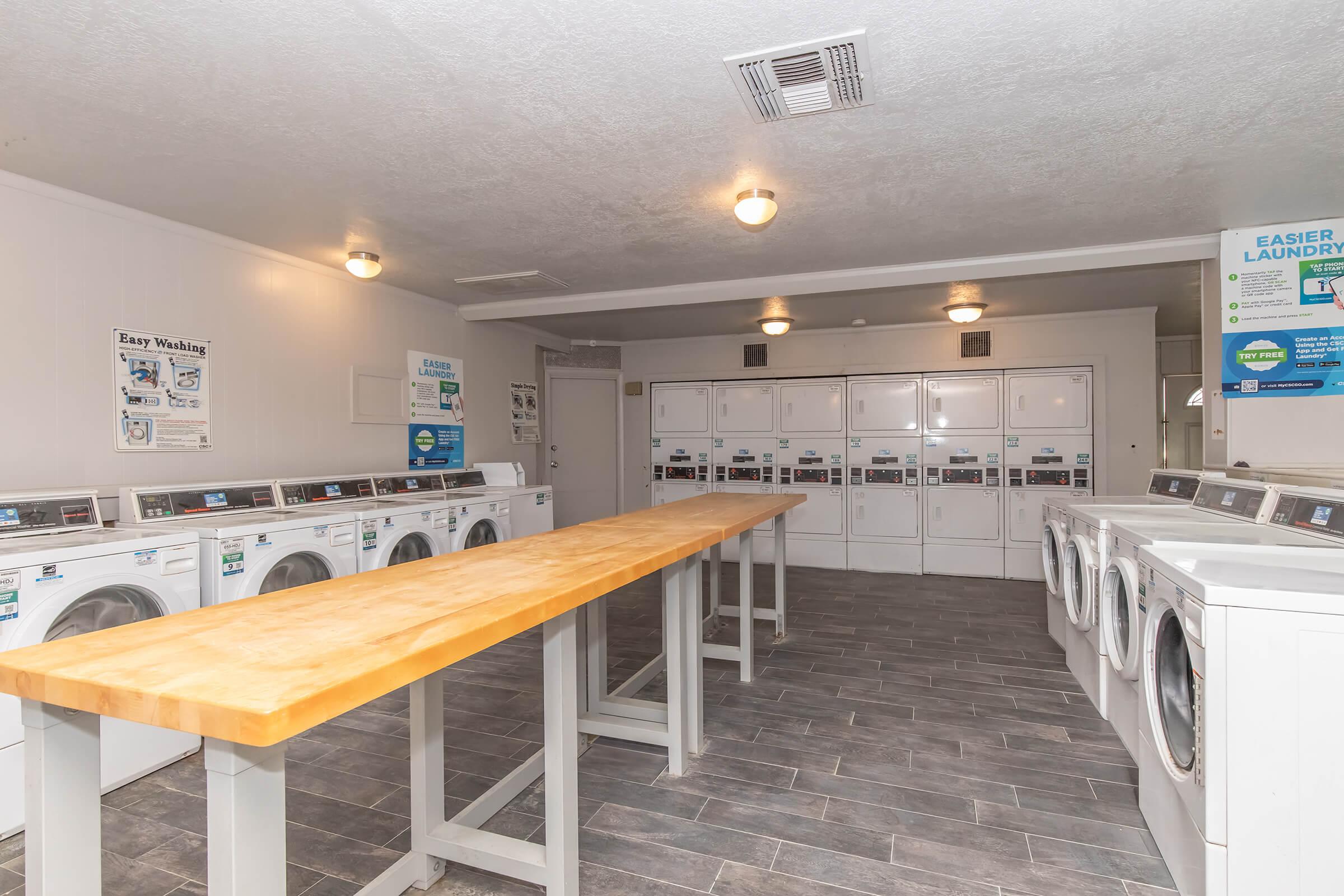 A modern laundry room featuring several washing machines and dryers lined against the walls. There is a long wooden table in the center, with bright overhead lighting. The walls are a neutral color, and there are informational posters about laundry services.