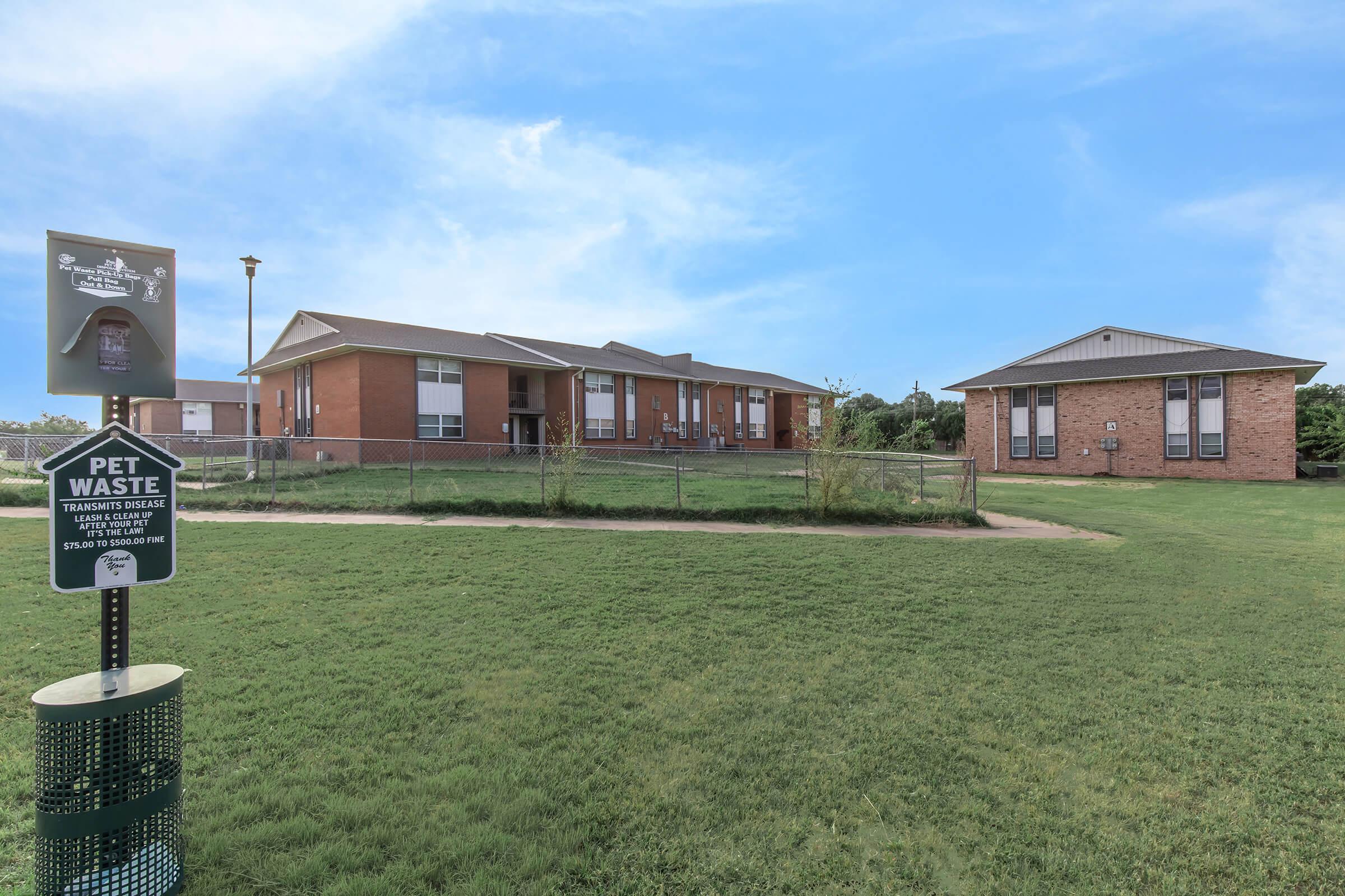 A grassy area with a "Pet Waste" sign and a waste disposal bin. In the background, there are two buildings: a single-story brick structure and another larger building. The sky is clear with a few clouds, creating a bright and open atmosphere. The area appears well-maintained.