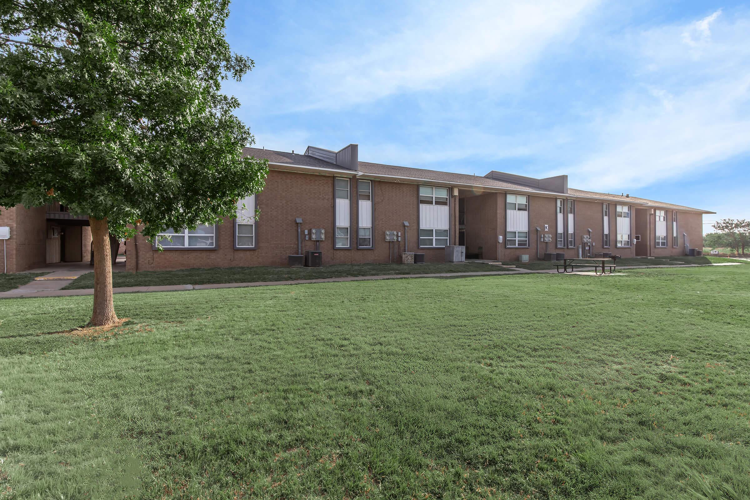 A single-story brick building with multiple windows, surrounded by green grass and a tree in the foreground. There are air conditioning units visible on the exterior walls, and a picnic table can be seen on the lawn. The sky is clear with some clouds.
