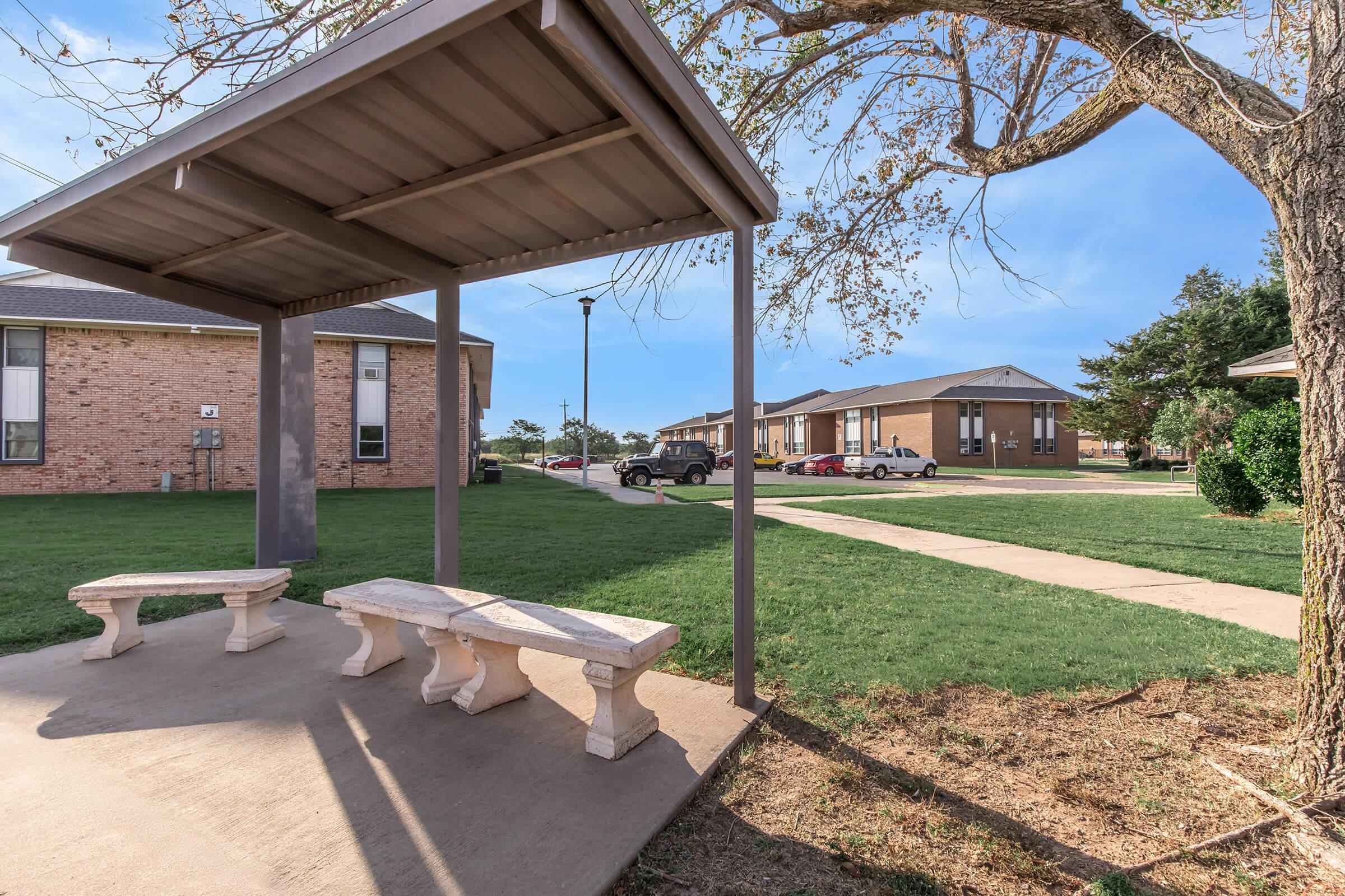 A shaded bus stop with stone benches in a grassy area, surrounded by residential buildings. A tree with sparse leaves is nearby, and a pathway leads to additional housing. Several parked cars are visible in the background under a clear blue sky.