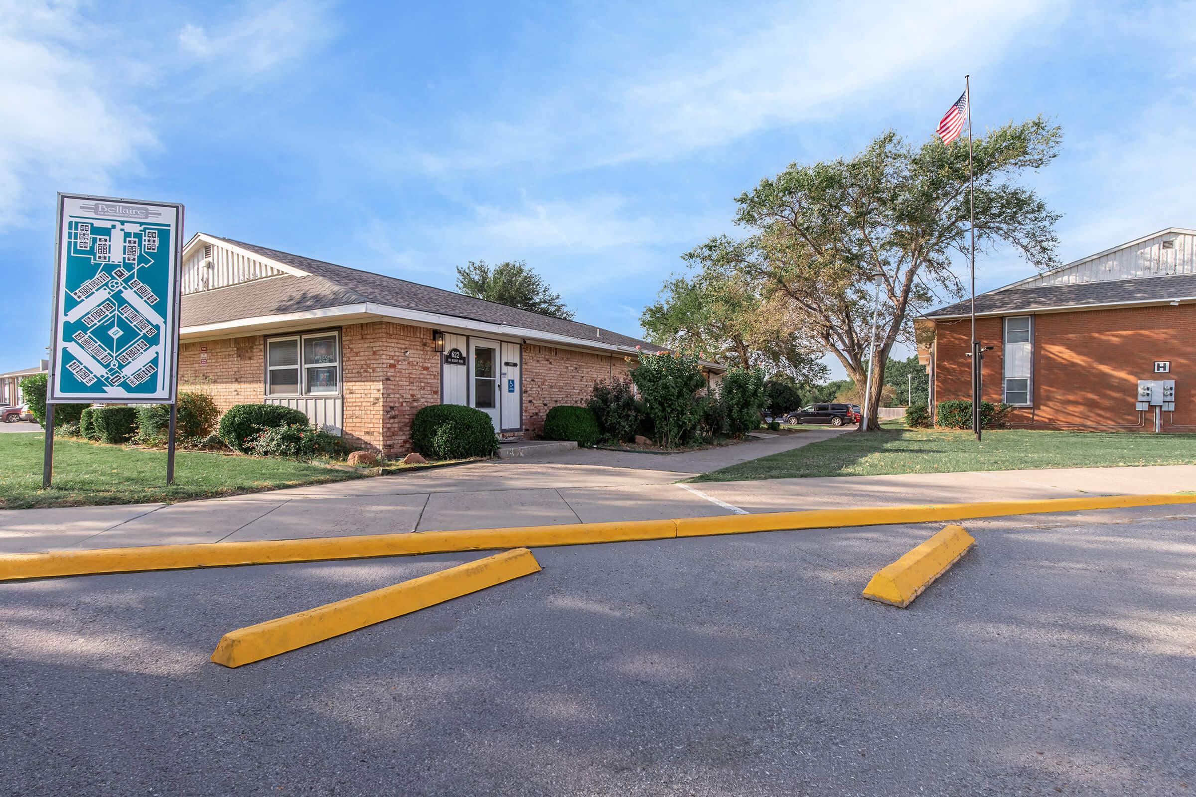 Brick building with a sloped roof and a sign displaying a map of the area. Lush green landscaping surrounds the building, which is situated near a parking lot with yellow curbs. A flagpole with an American flag stands nearby against a clear blue sky. Trees provide additional greenery in the background.