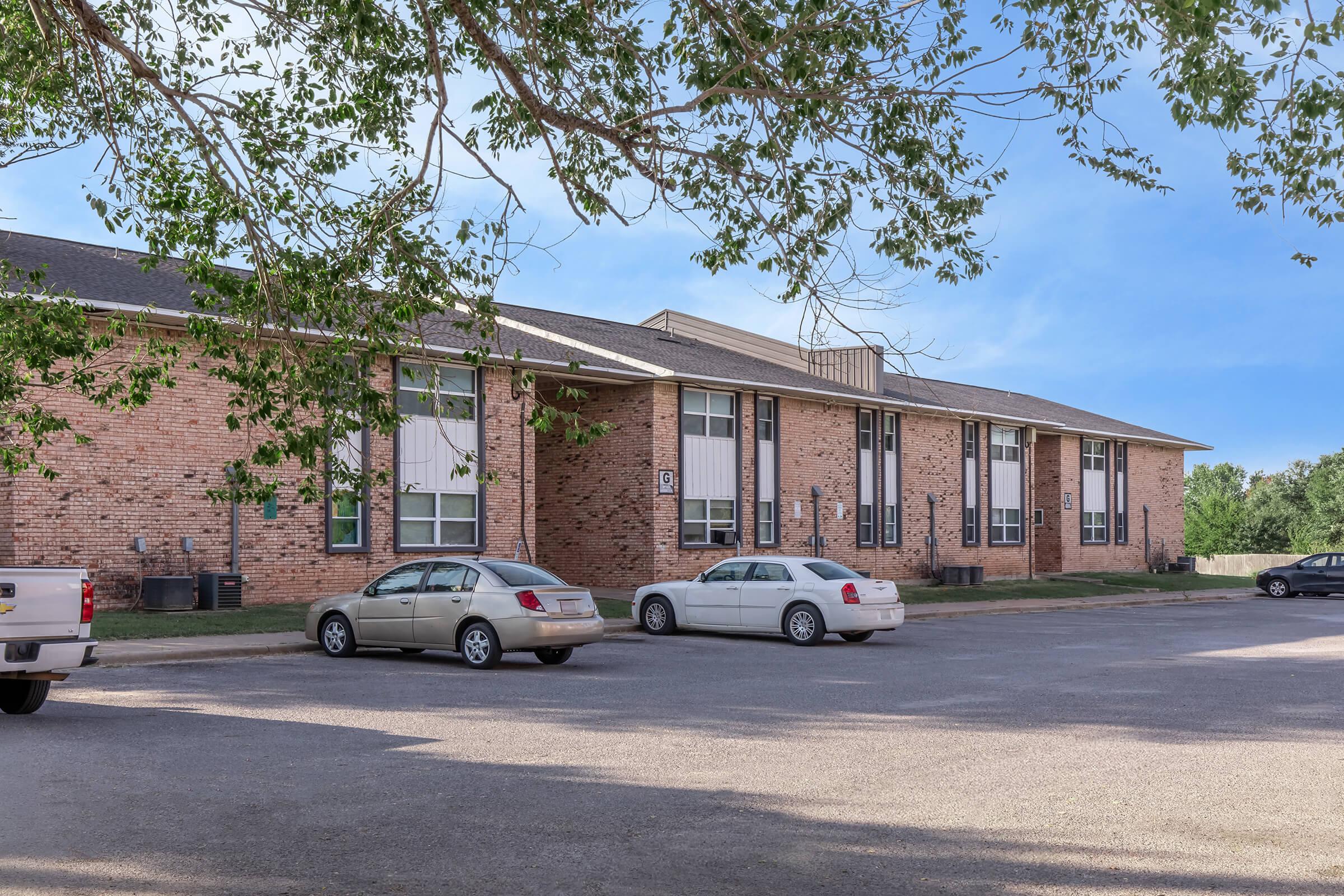 A brick building with multiple windows and a flat roof is shown. There is a gravel parking lot in front with several parked cars. Trees are visible on the left side, providing shade. The sky is clear and blue, indicating a sunny day.