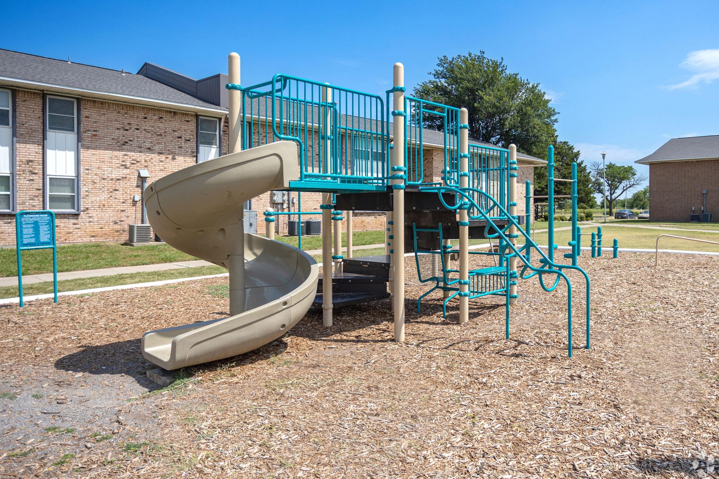 A playground structure featuring a large slide, climbing features, and multiple levels, situated on a bed of wood chips. The background includes brick buildings and green grass under a bright blue sky.