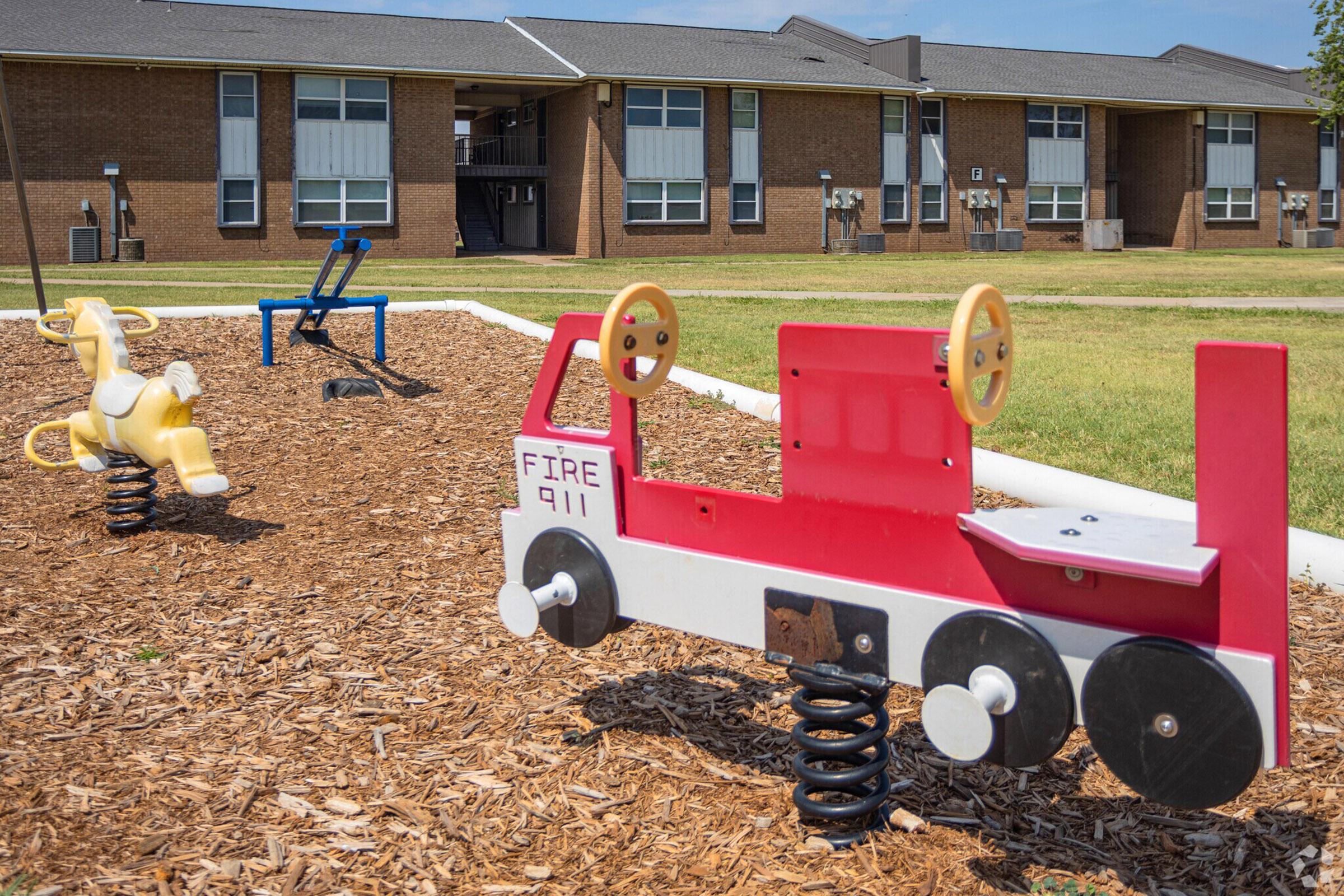Playground area with spring-mounted toys including a red fire truck and a yellow animal. The background features brick buildings and a grassy lawn under a clear sky, creating a vibrant outdoor space for children.