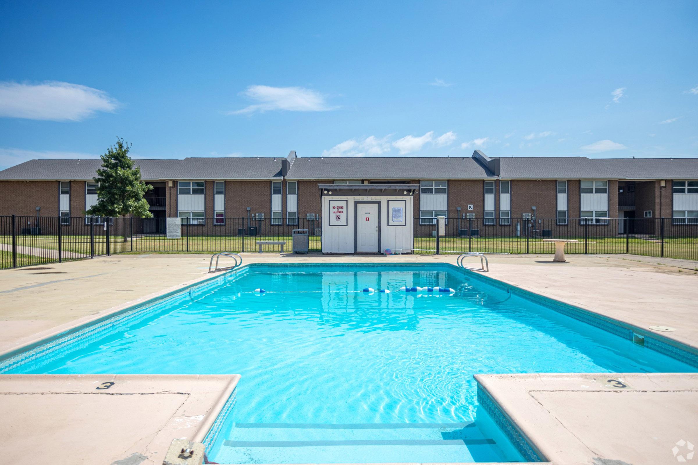 A clear blue swimming pool with steps leading into it, surrounded by a concrete deck. In the background, there are two-story apartment buildings with grass and a tree. The sky is bright blue with a few clouds, creating a sunny atmosphere. A fence encloses the pool area.