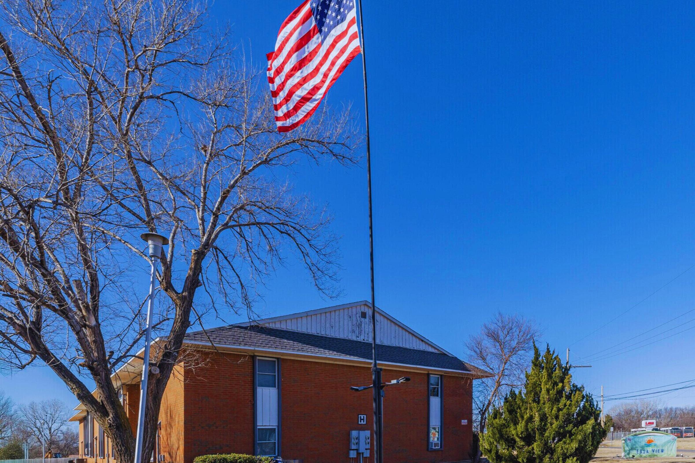 A clear blue sky serves as the backdrop for a large American flag flying on a tall pole in front of a brick building. To the right, a green bush and a tree are visible, adding to the scene's natural elements. The building appears to be a modest structure in a suburban area.