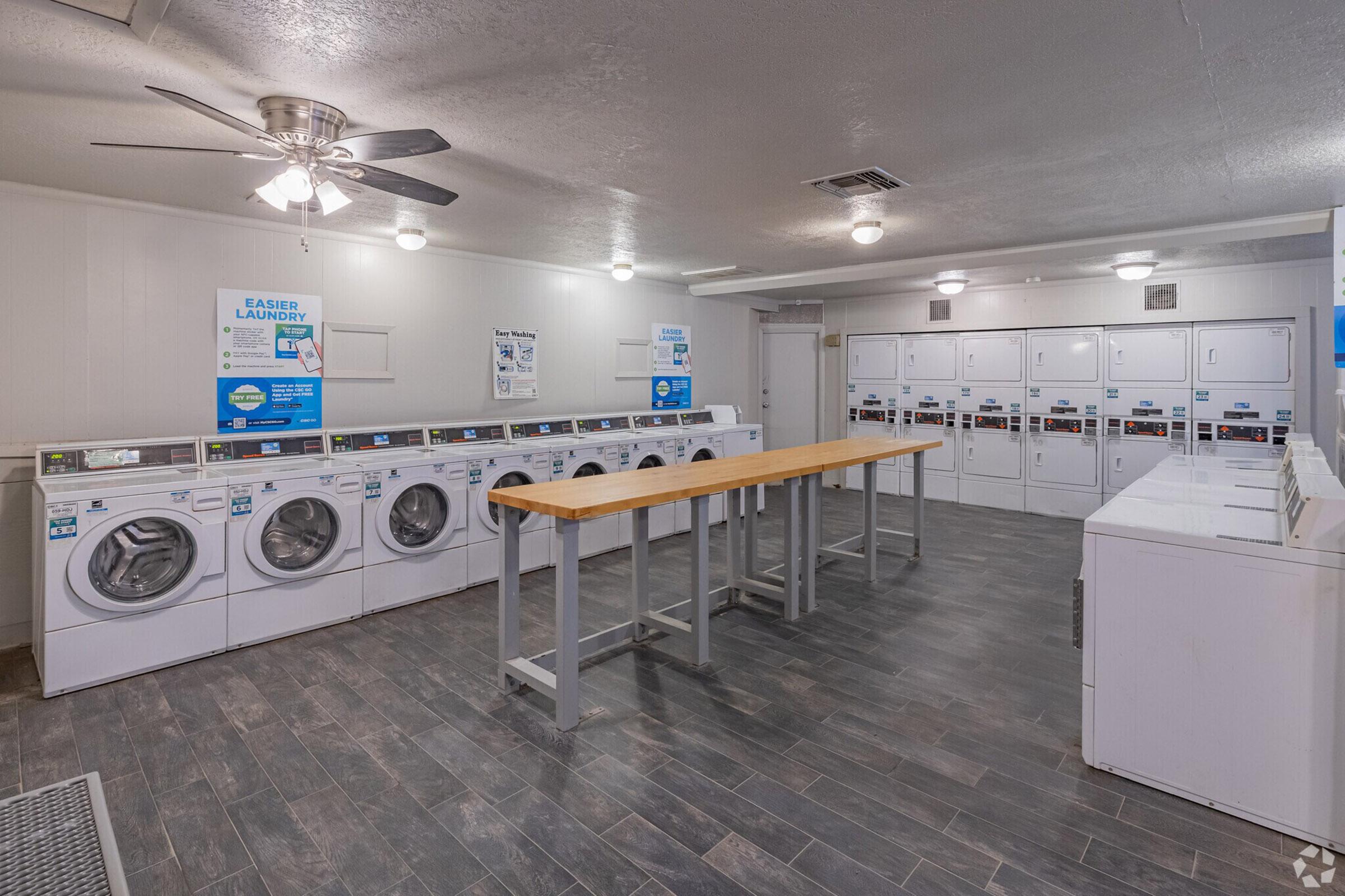 A clean, well-lit laundry room featuring multiple washing machines and dryers lined against the walls, along with a central folding table. The space has modern decor, gray tiled flooring, and information posters on the walls, creating a functional and inviting environment for laundry tasks.