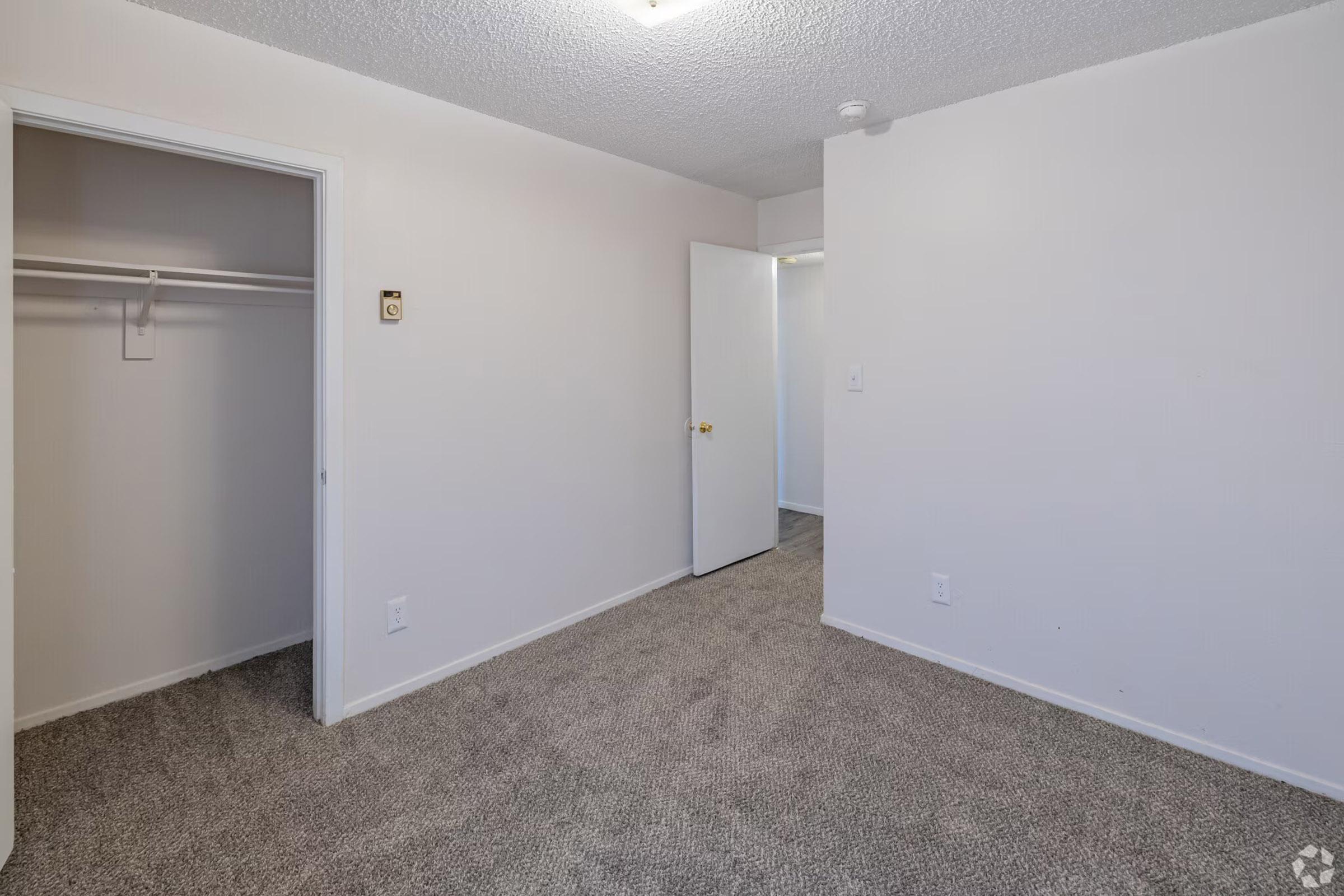 A vacant room featuring beige carpet flooring, light-colored walls, and a closet with a sliding door. There's a door leading to another room in the background, and a ceiling light fixture is visible. The space is well-lit and clean, providing a blank canvas for potential decoration.