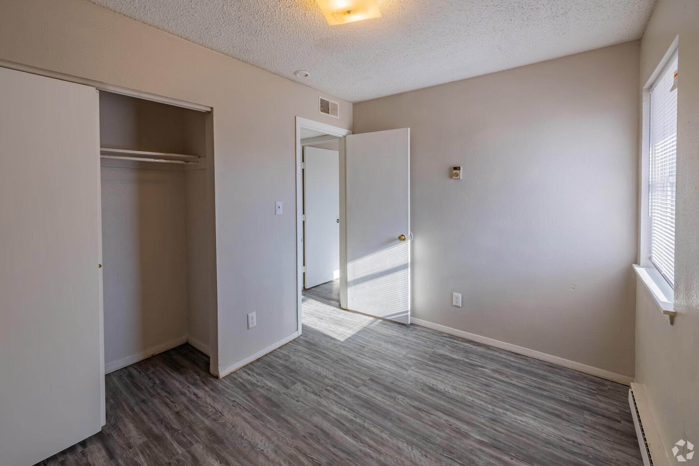A well-lit empty room with light gray walls and a laminate floor. There is a closet on the left, a door leading to another area on the right, and a window allowing natural light in. The room has a simple design, suitable for various purposes such as a bedroom or office space.