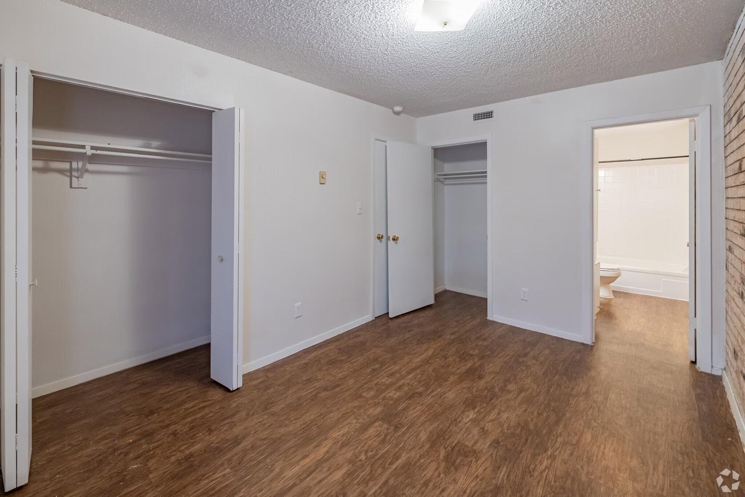 A spacious, well-lit room with wood laminate flooring. Features two closet spaces with sliding doors on the left, and a bathroom doorway visible at the back, along with a plain white wall and textured ceiling.