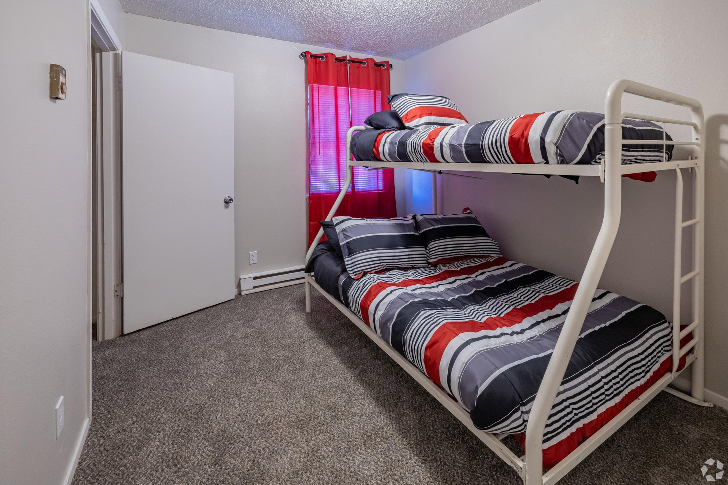 A cozy bedroom featuring a bunk bed with striped bedding in red, gray, and white. The room has a window dressed with red curtains, beige walls, and carpet flooring. A closed white door is visible on the left side, adding to the welcoming atmosphere of the space.
