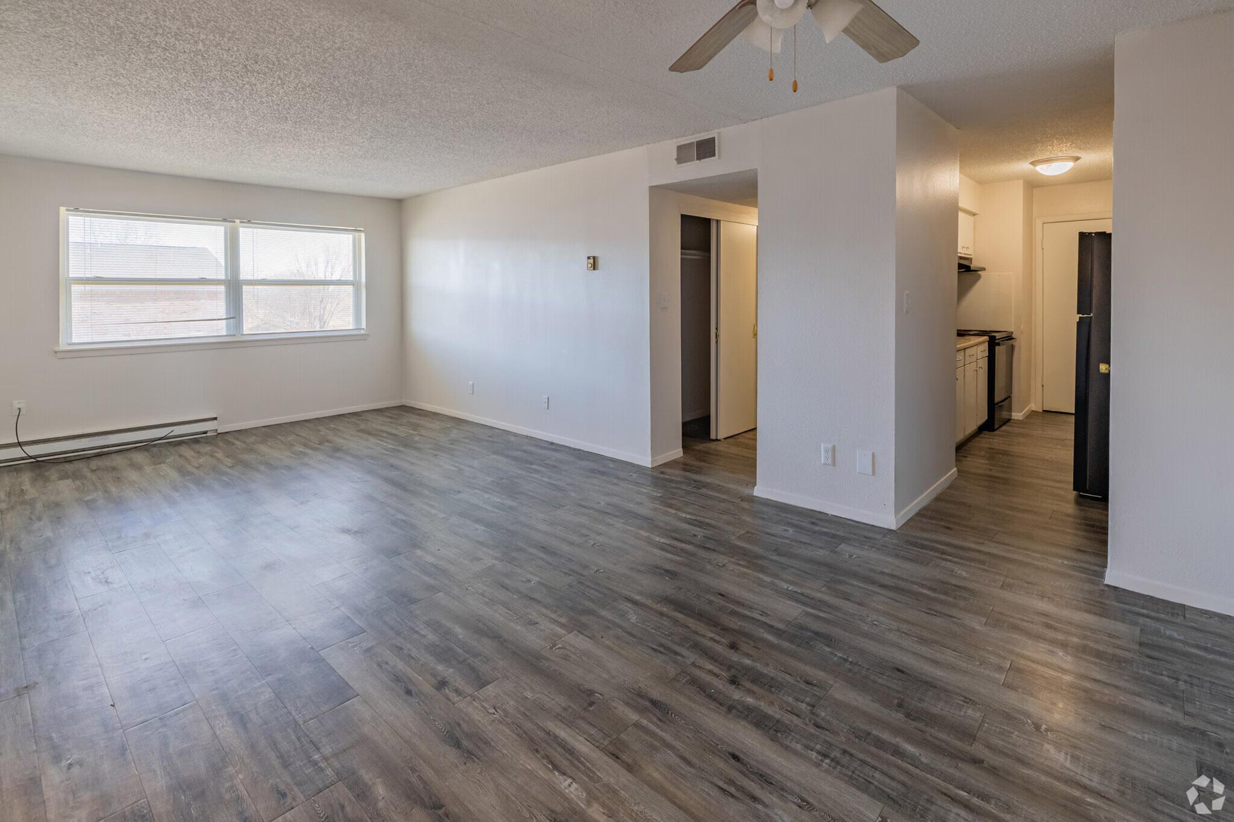 A spacious, empty living area with light-colored walls and laminate flooring. A ceiling fan is visible, and there are windows allowing natural light. To the right, a doorway leads to a kitchen area, with appliances and cabinets in view. The overall vibe is modern and bright.
