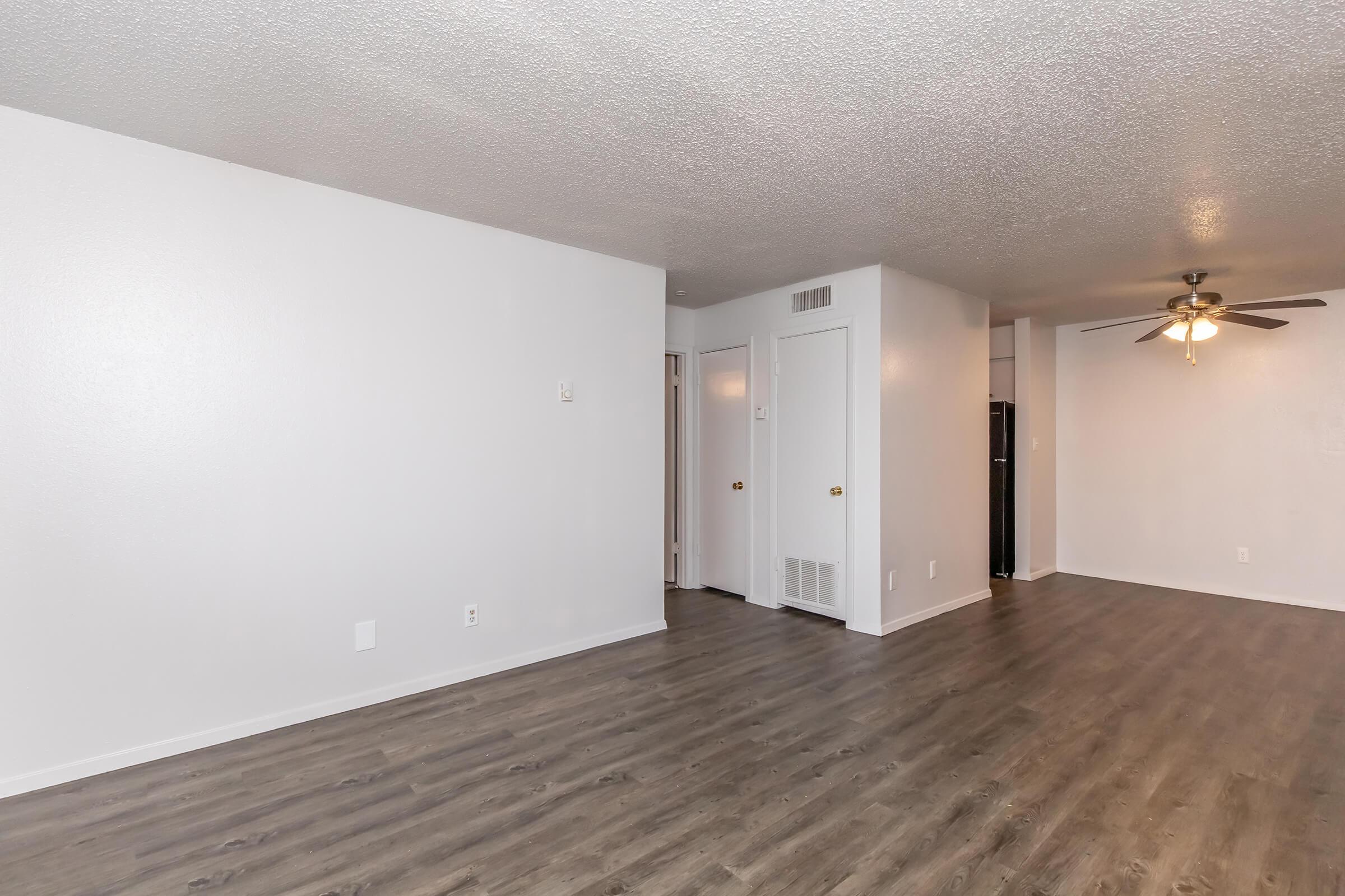 Bright, open living space featuring light-colored walls, laminate flooring, and a ceiling fan. The room includes a closed door leading to another room and an open entryway, with a glimpse of a kitchen area in the background. Ideal for showcasing a modern, comfortable apartment layout.