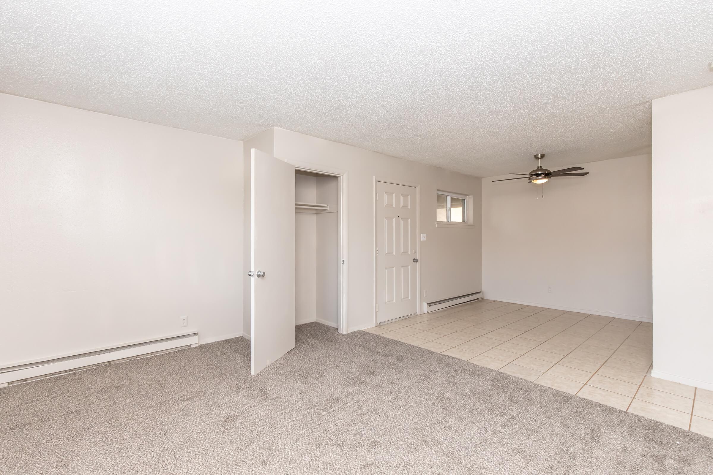A spacious, empty living room featuring light gray carpet and beige walls. There is a closet with a white door on the left, and a second door leading to another room. A ceiling fan hangs in the center, and the floor is tiled near the entrance. Natural light enters through a small window.
