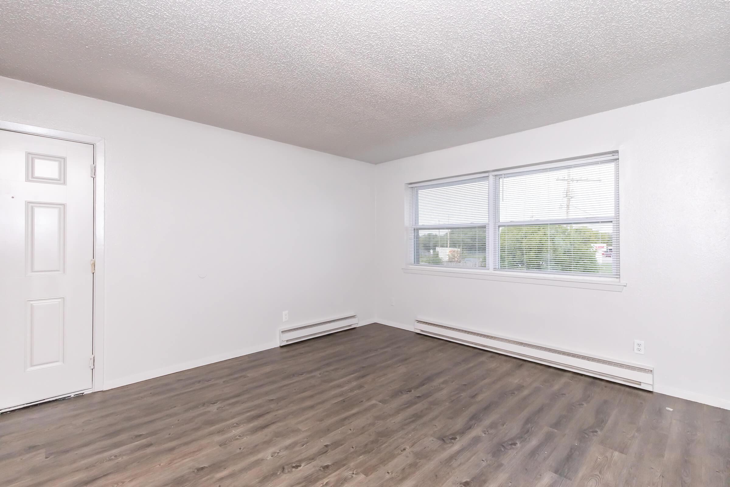 Empty room featuring light-colored walls and a textured ceiling. Natural light comes in through a large window with blinds. The floor is covered in a warm-toned wood laminate. A front door is visible on the left side, and there are baseboard heaters along the walls.